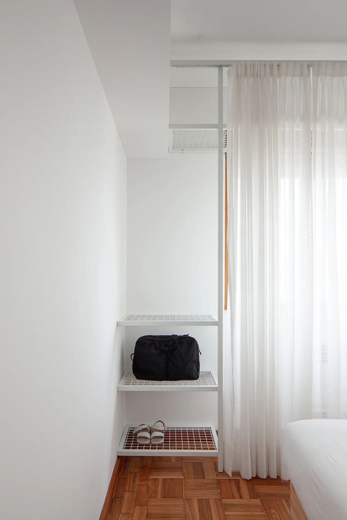 A white metal wire shelving unit holds a black bag and sandals in a bedroom corner next to sheer curtains and a wood parquet floor.