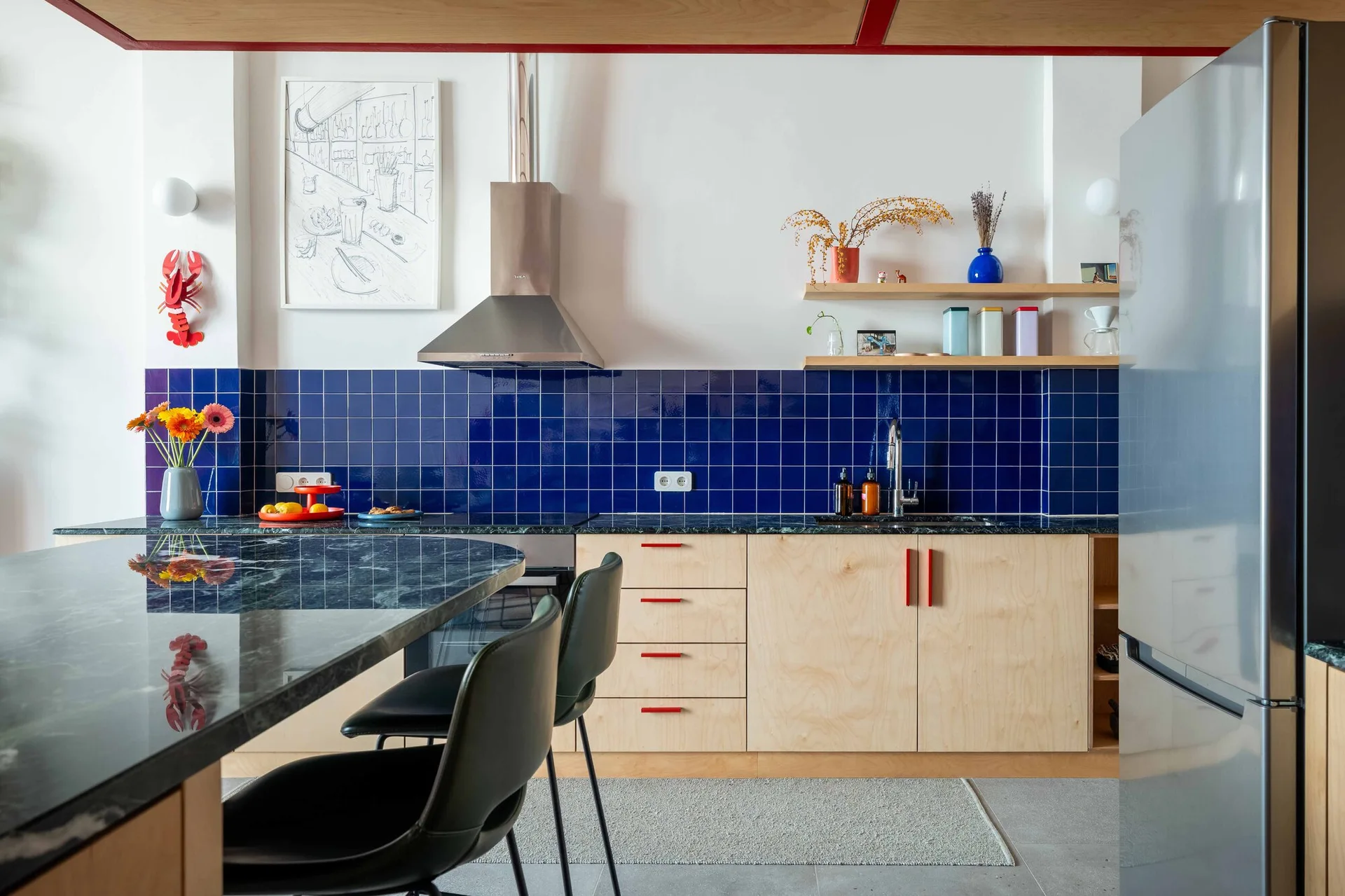 A modern kitchen featuring light plywood cabinets with red handles, a deep blue tiled backsplash, and a dark marble kitchen island.