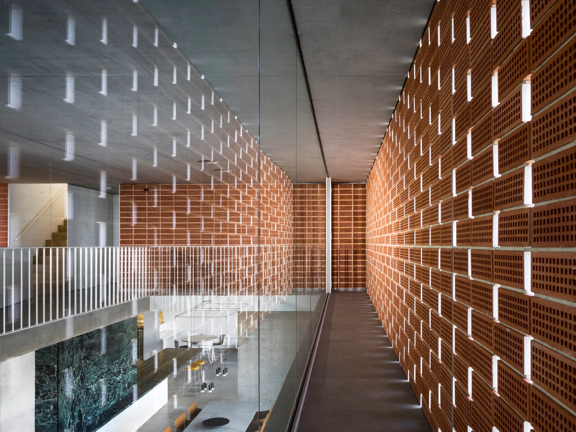 An elevated concrete walkway features a full-height glass partition and a patterned terracotta brick lattice wall overlooking a lower level.