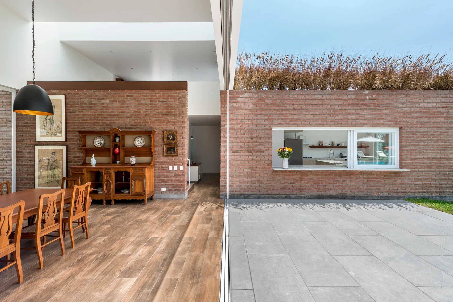 A dining room with wood flooring and a brick wall transitions seamlessly onto a stone patio with a pass-through serving window.