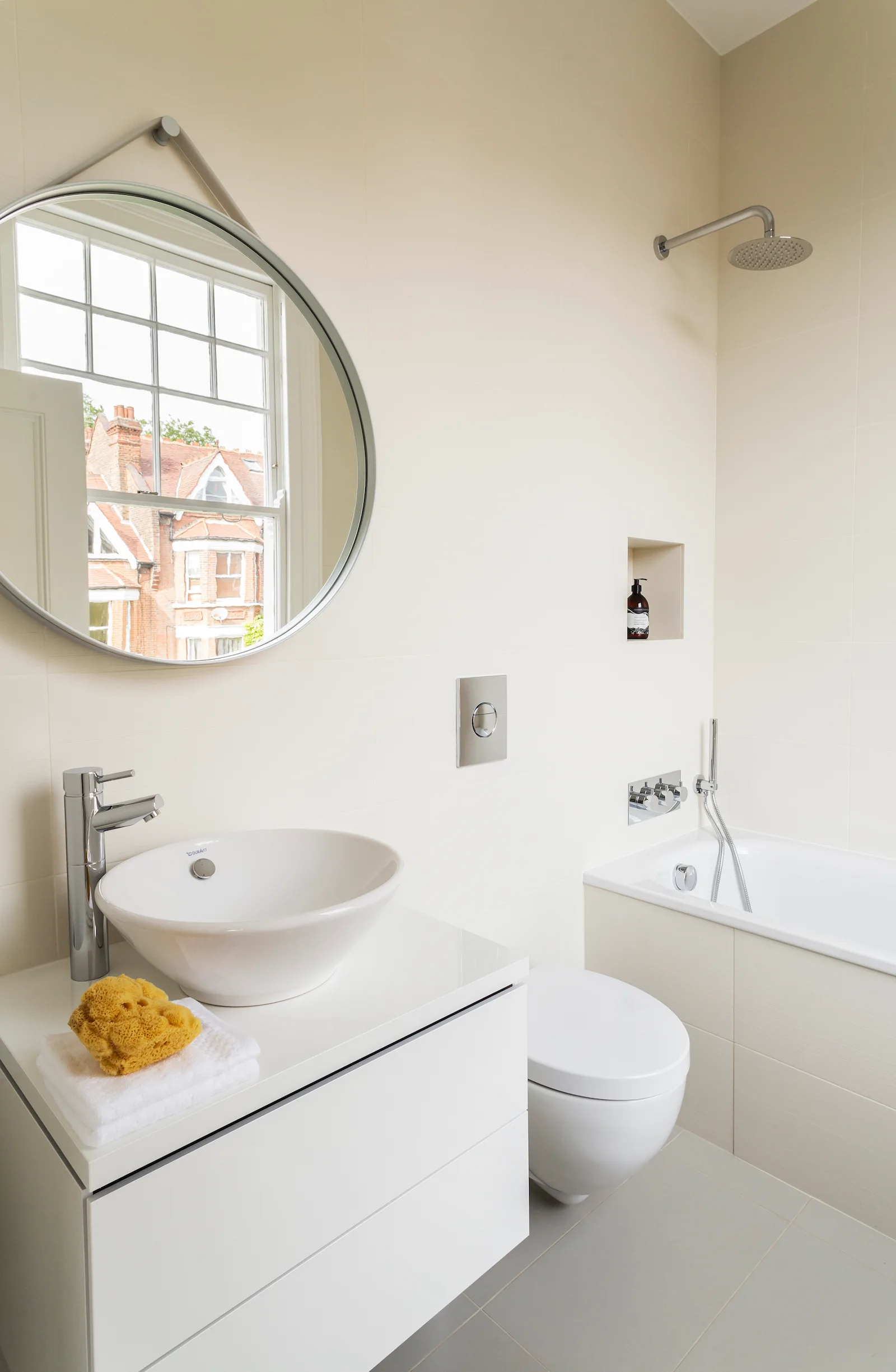 A modern cream-tiled bathroom featuring a white vanity with a vessel sink, a wall-mounted toilet, and a built-in bathtub.