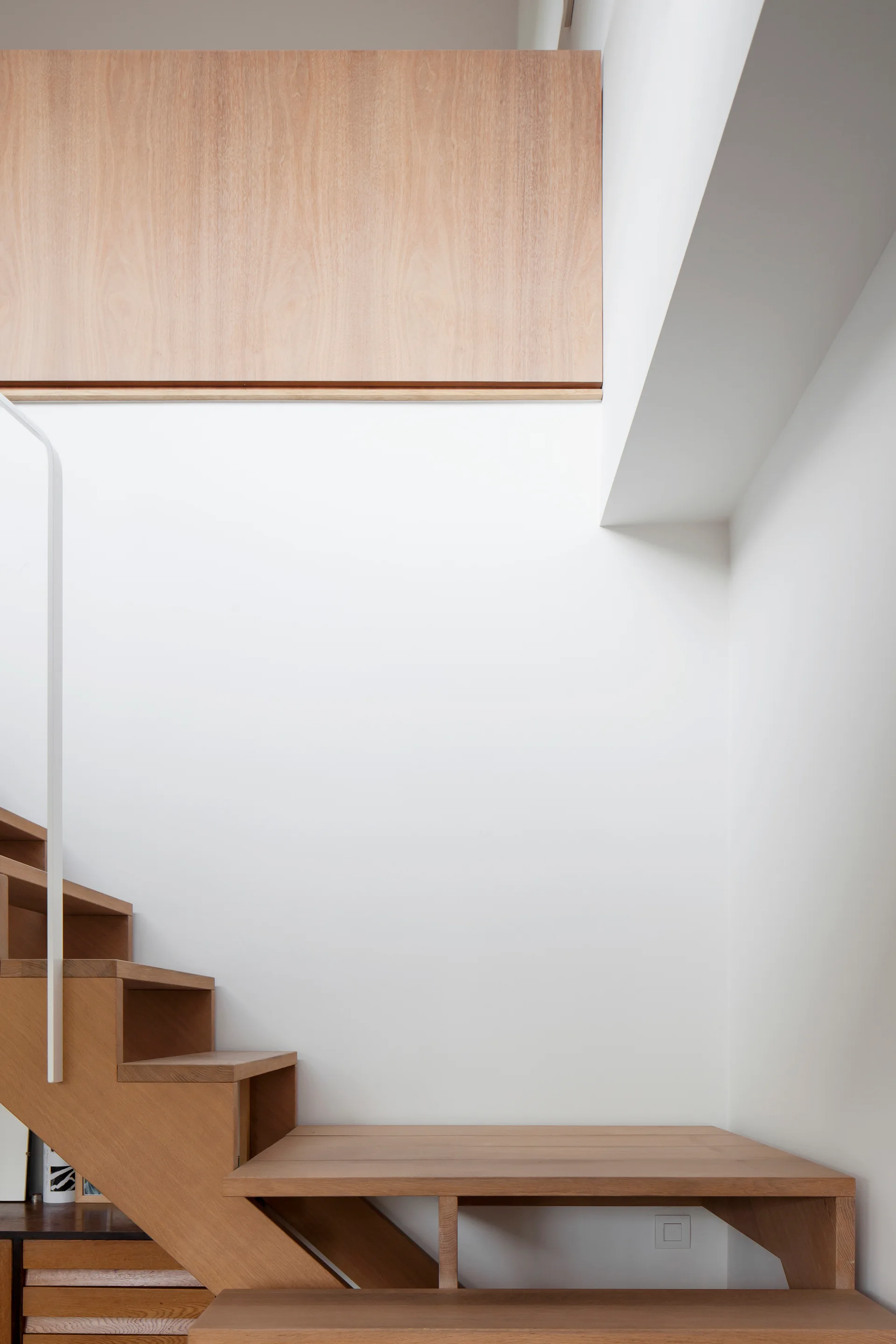 A minimalist wooden staircase with light-colored treads features a thin white metal railing against a solid white wall.