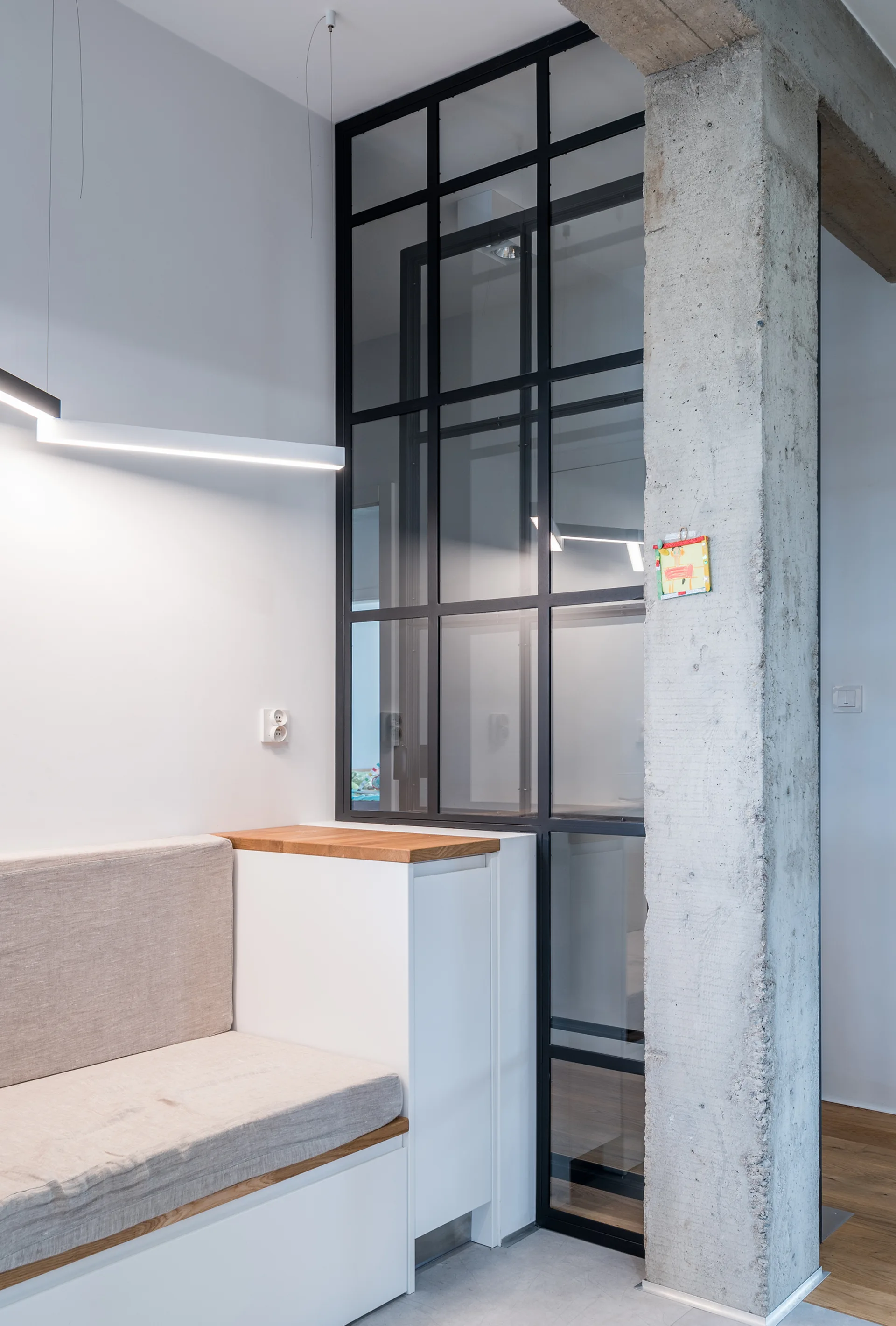 A modern interior features a built-in bench with a beige cushion next to an exposed concrete column and a black metal-framed glass partition.