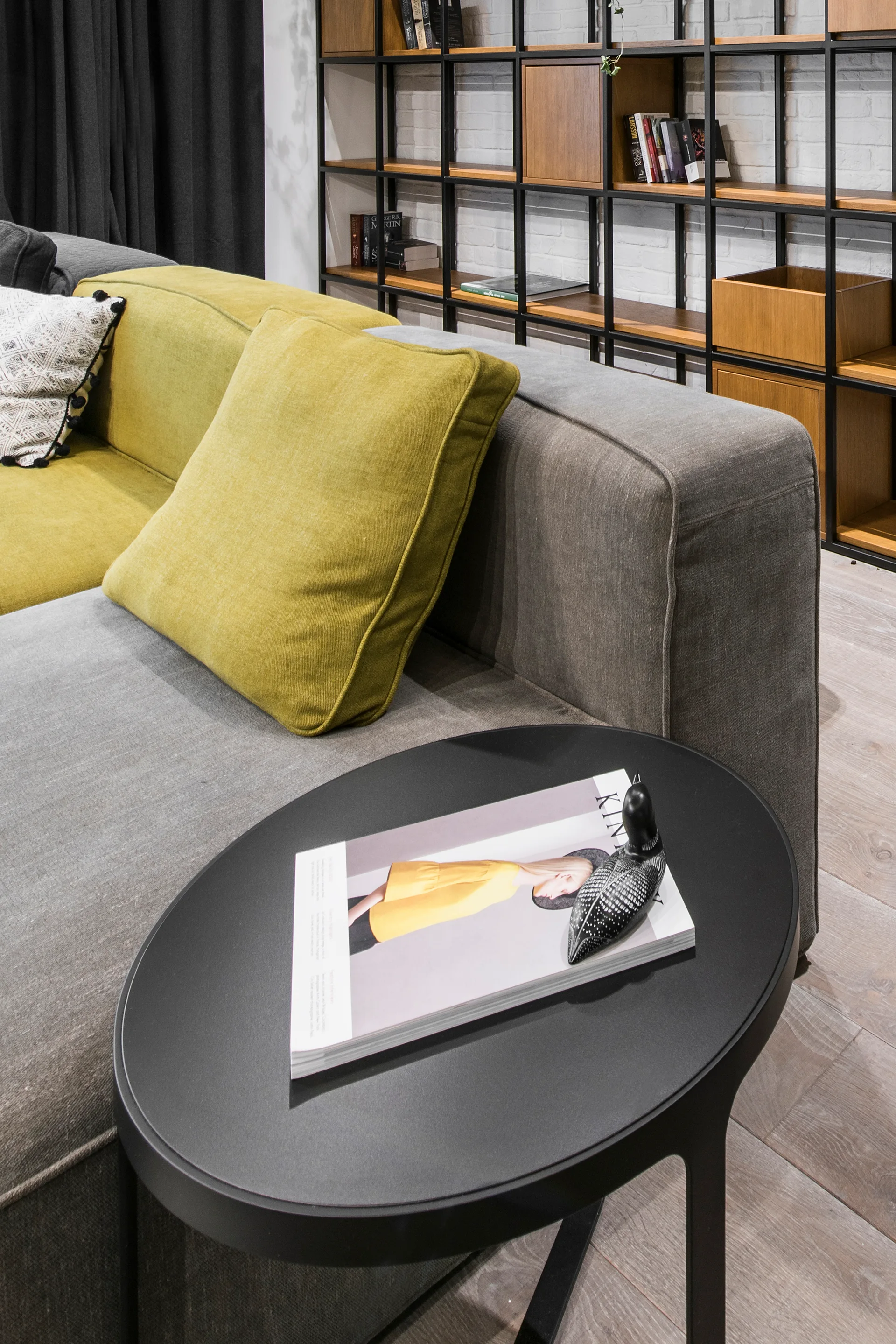 A grey modular sectional sofa with a lime green cushion sits next to a black round side table and a tall black metal grid bookcase.