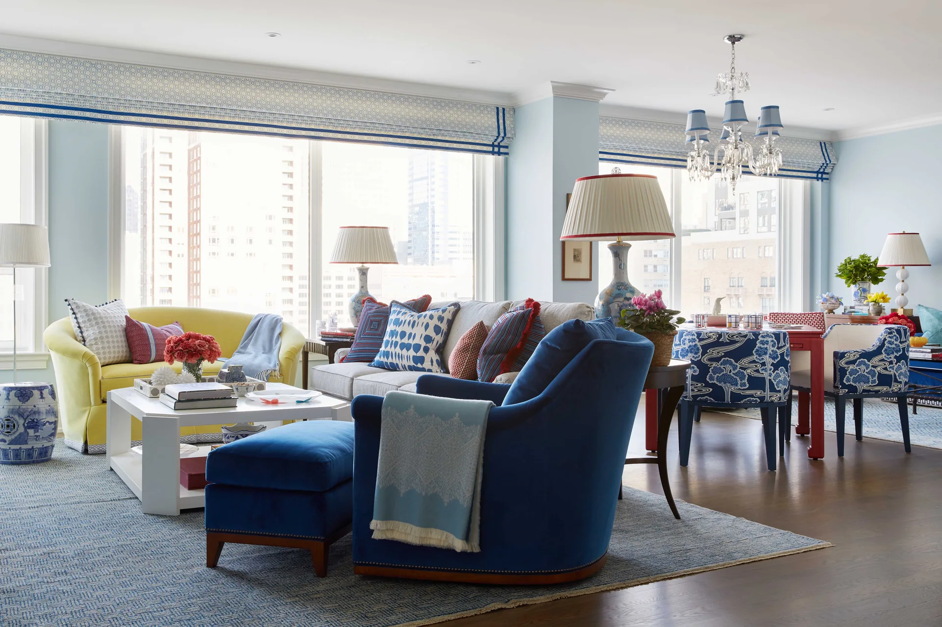 An eclectic living room features a blue velvet armchair, a yellow sofa, and a dining area with patterned chairs under a crystal chandelier.