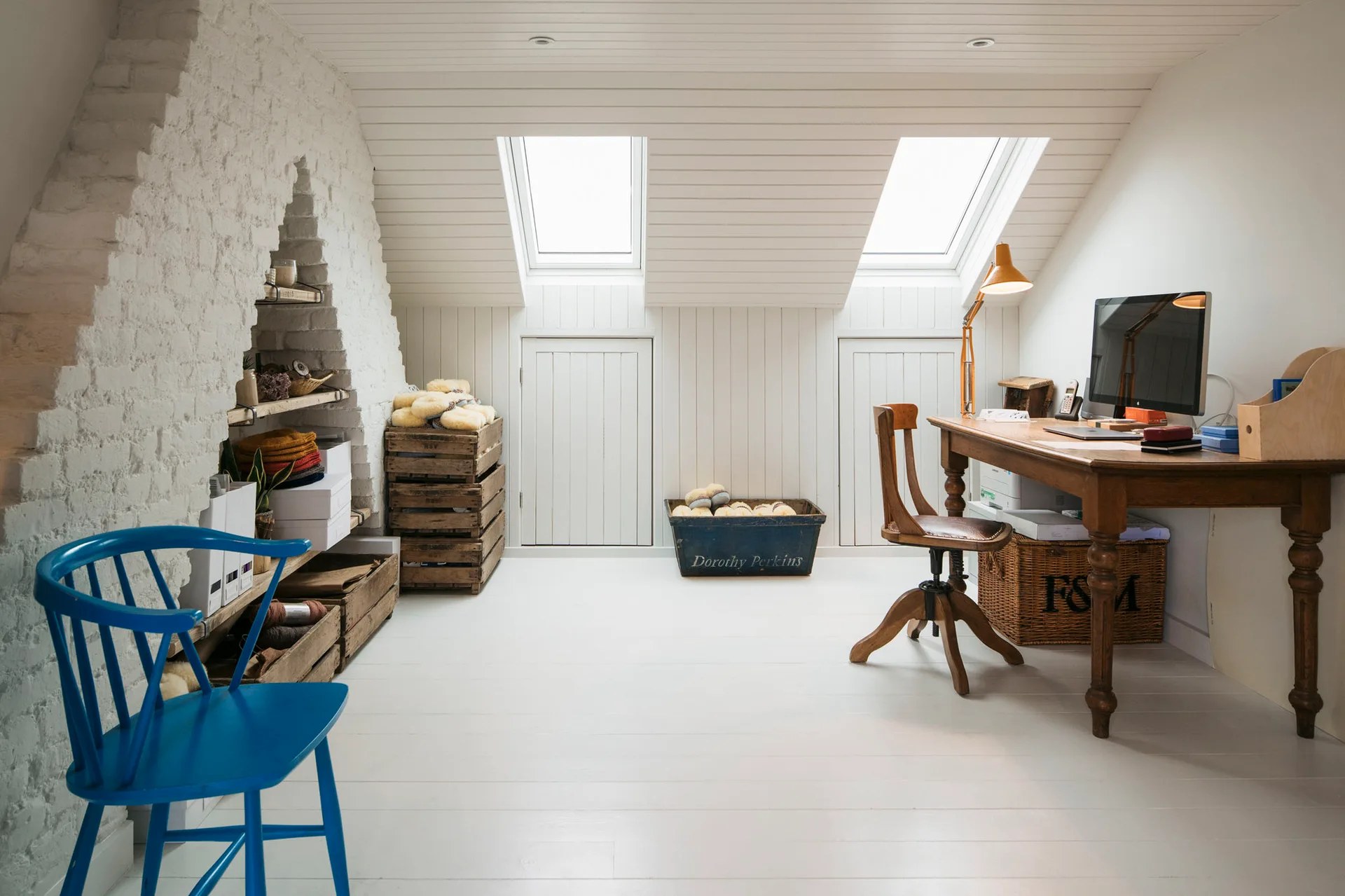 A bright attic home office features a sloped ceiling with two skylights, an exposed white brick wall with shelves, and a wooden writing desk.