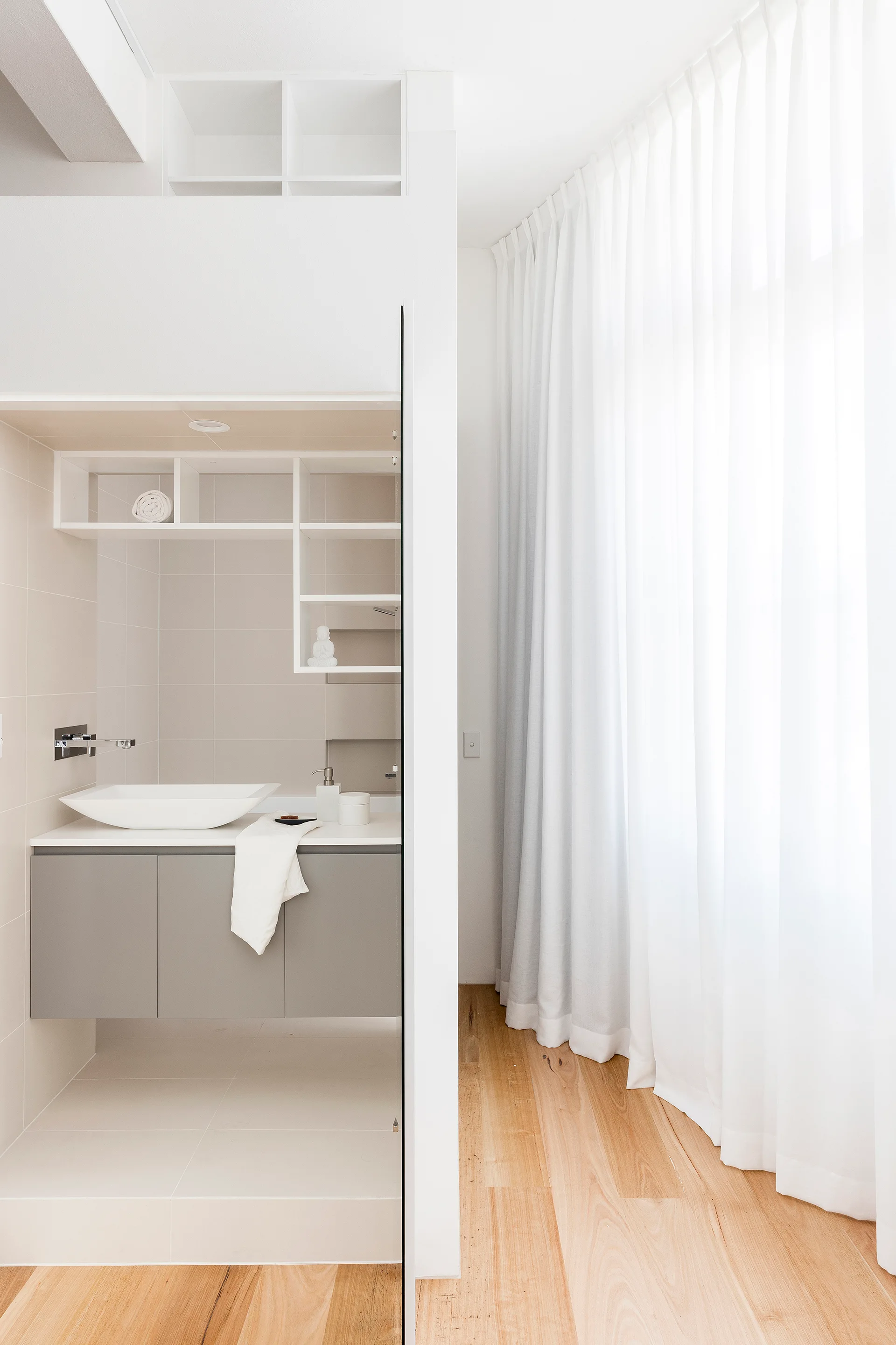A modern bathroom niche features a floating grey vanity with a white vessel sink, set beside a bright room with sheer white floor-length curtains.