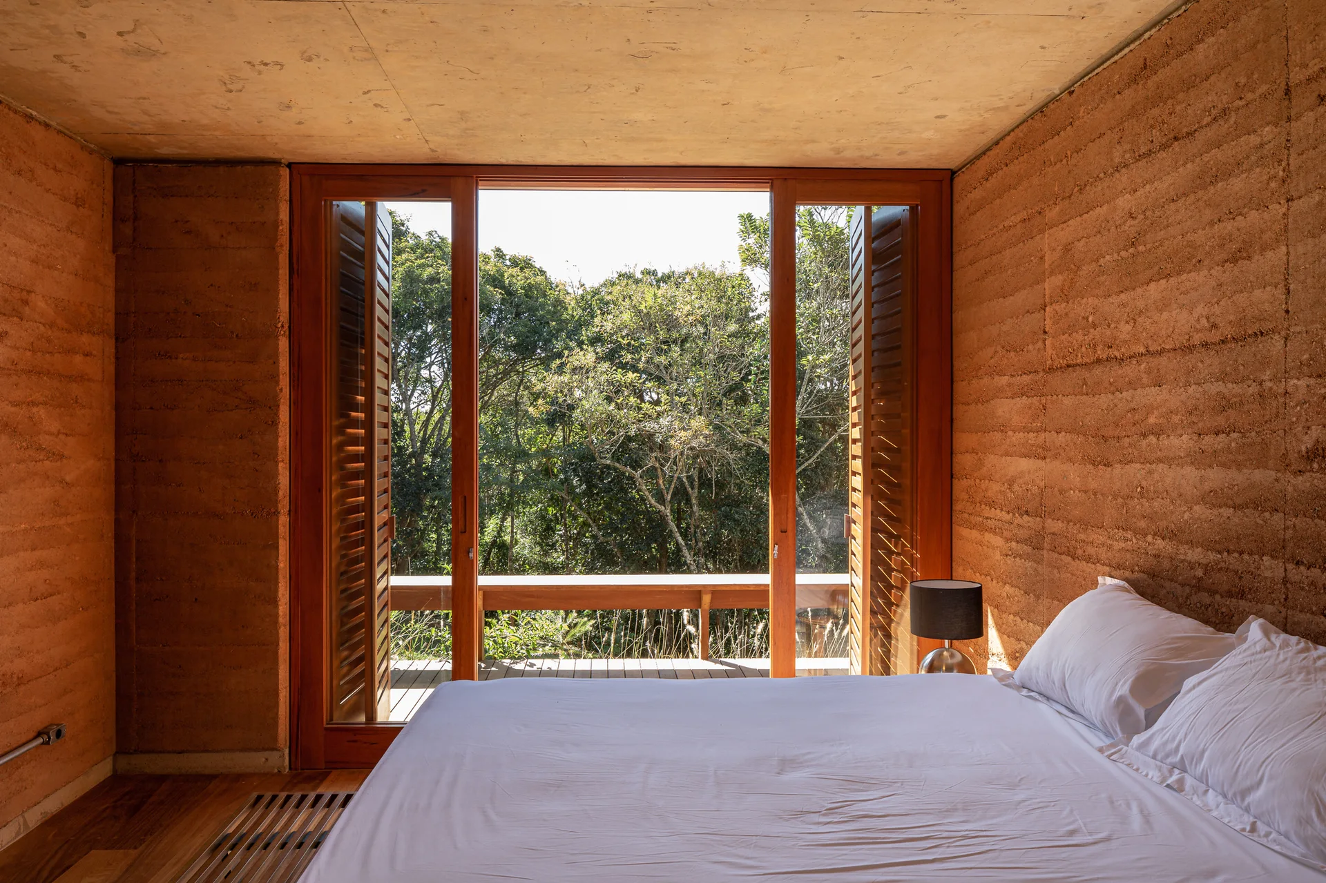 A bedroom with textured terracotta rammed earth walls, wooden floors, and glass doors with louvered shutters opening to a forest view.