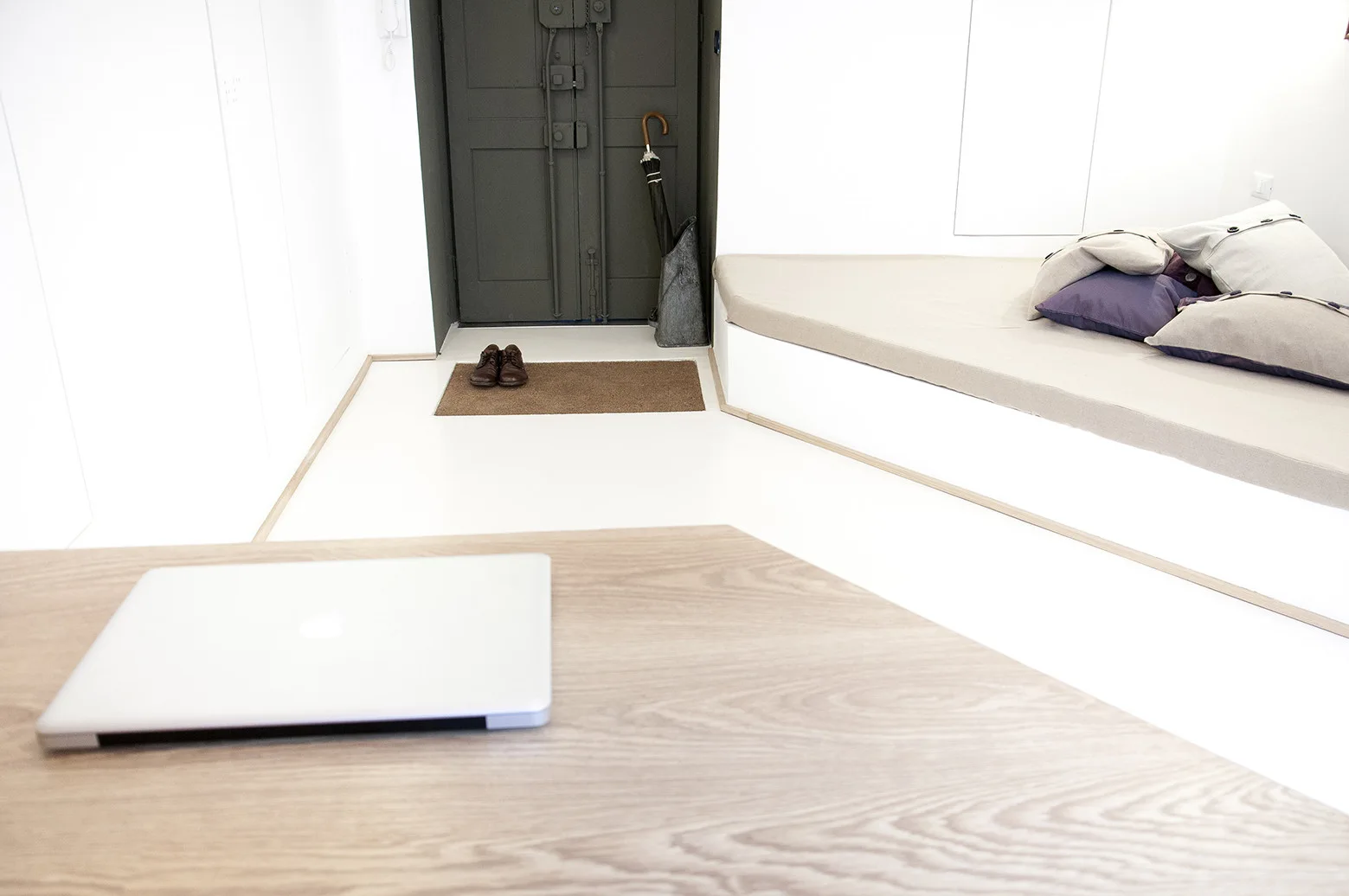 A bright minimalist entryway featuring a low-profile angular built-in bench with pillows, a dark front door, and a wooden table in the foreground.