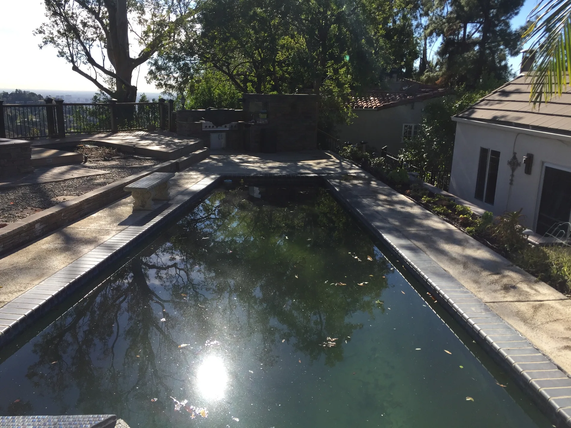 A rectangular swimming pool with dark grey tile coping is surrounded by a light concrete patio, a stone bench, and green trees.