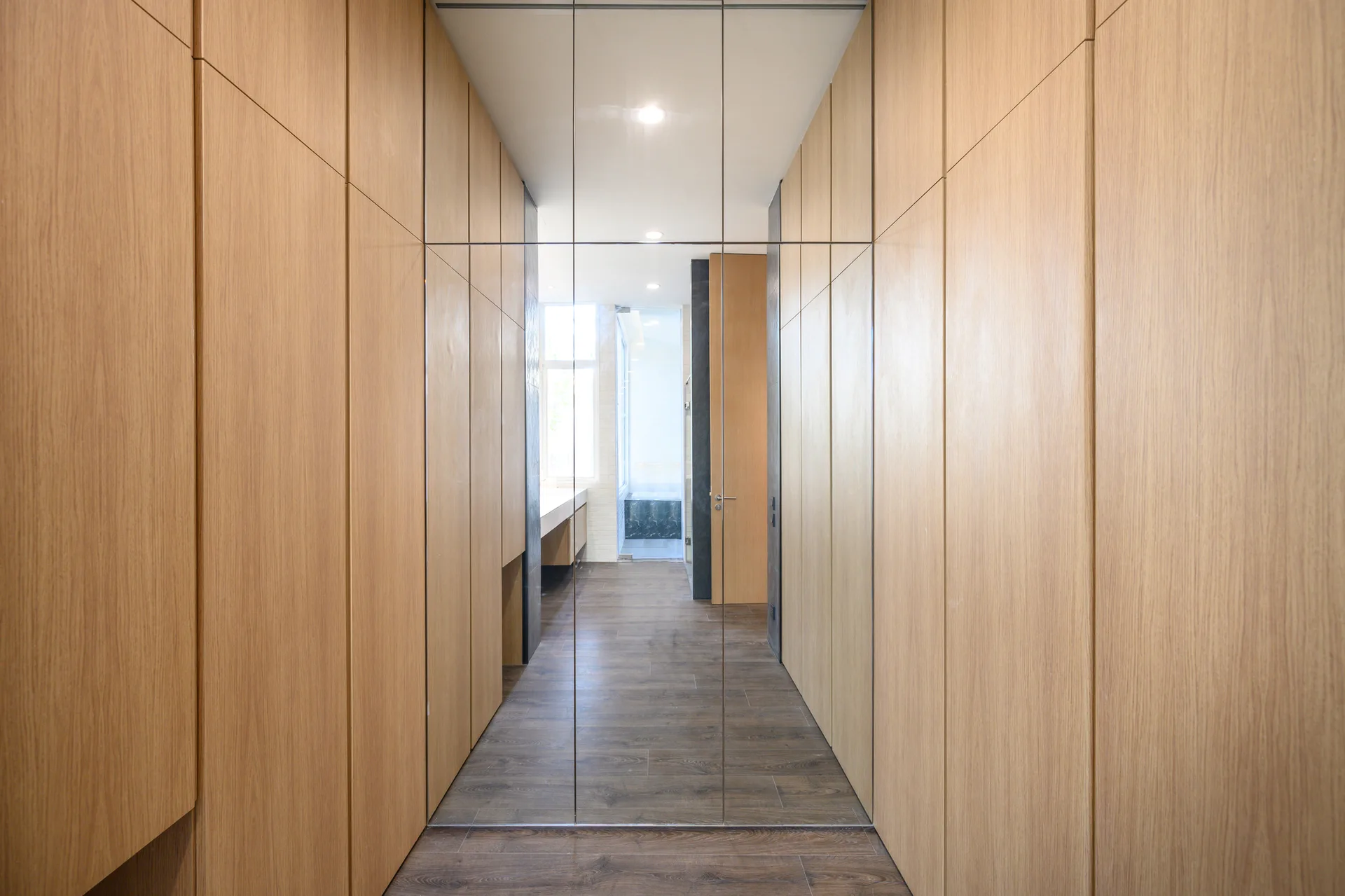 A narrow hallway lined with tall, light wood storage cabinets, featuring dark wood plank floors and a large floor-to-ceiling mirror.