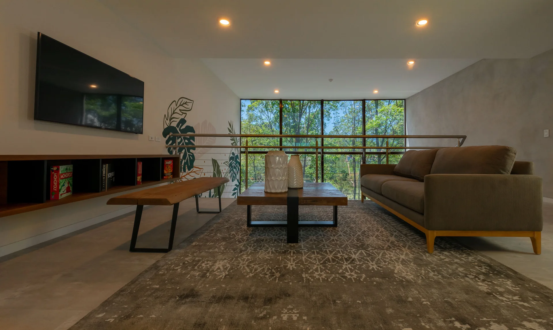 A modern loft living area features a gray sofa, a low wooden coffee table on a patterned rug, and a window overlooking trees.
