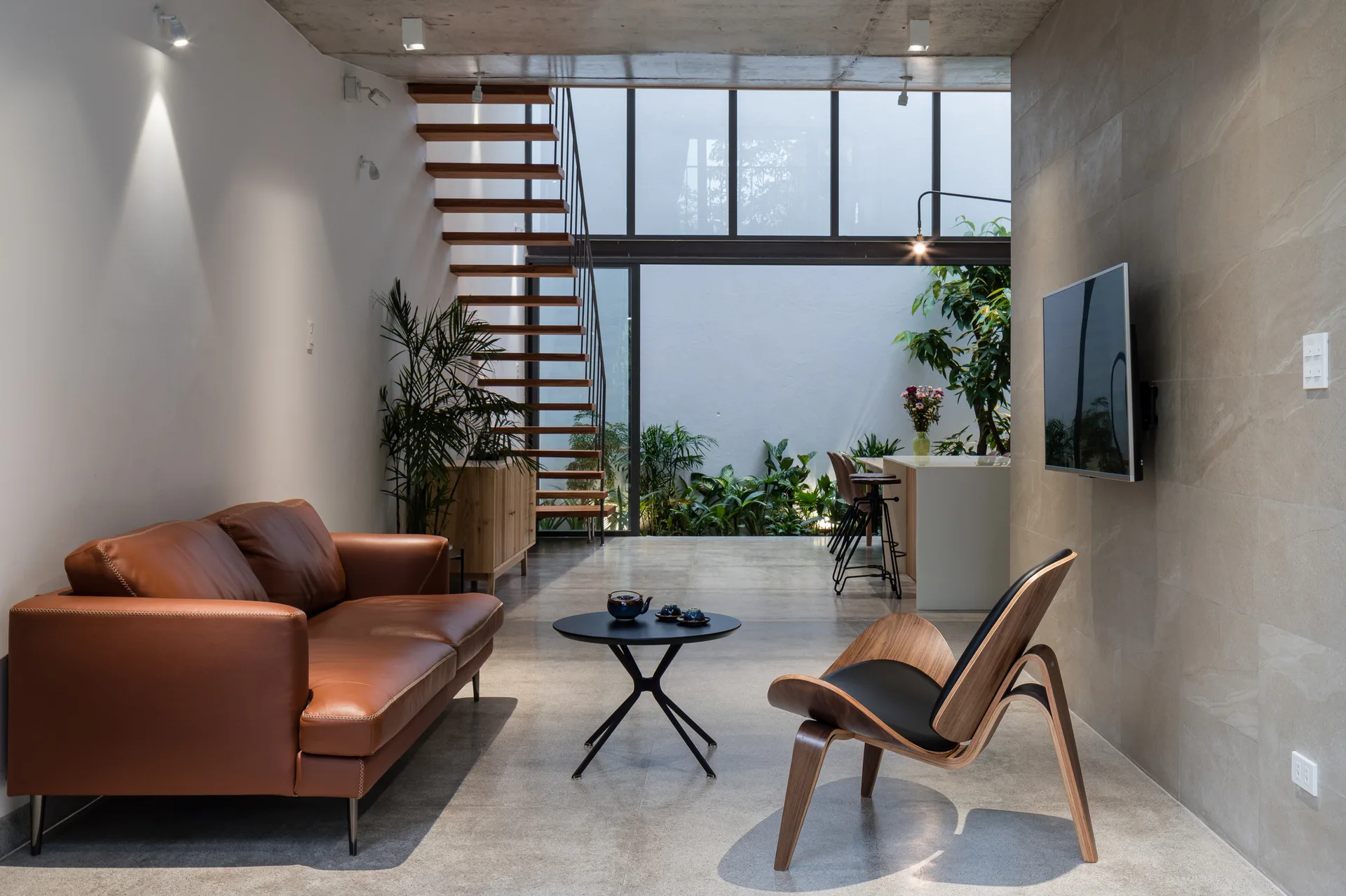A modern living room features a brown leather sofa, a curved wood lounge chair, and an open riser wooden staircase leading past a large window.