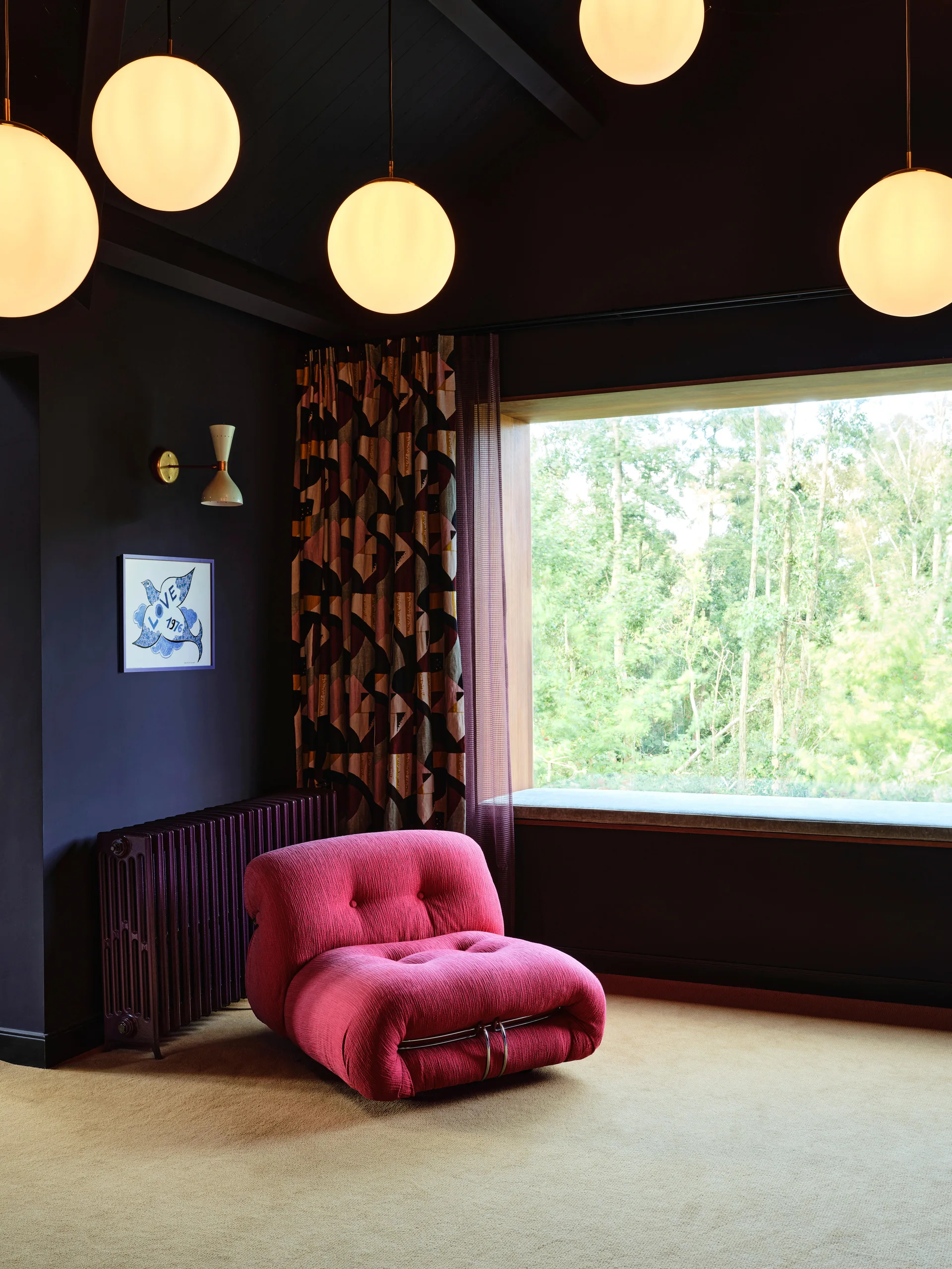 A tufted pink armchair sits on tan carpet in a dark charcoal room featuring globe pendant lights and a large window looking out at green trees.