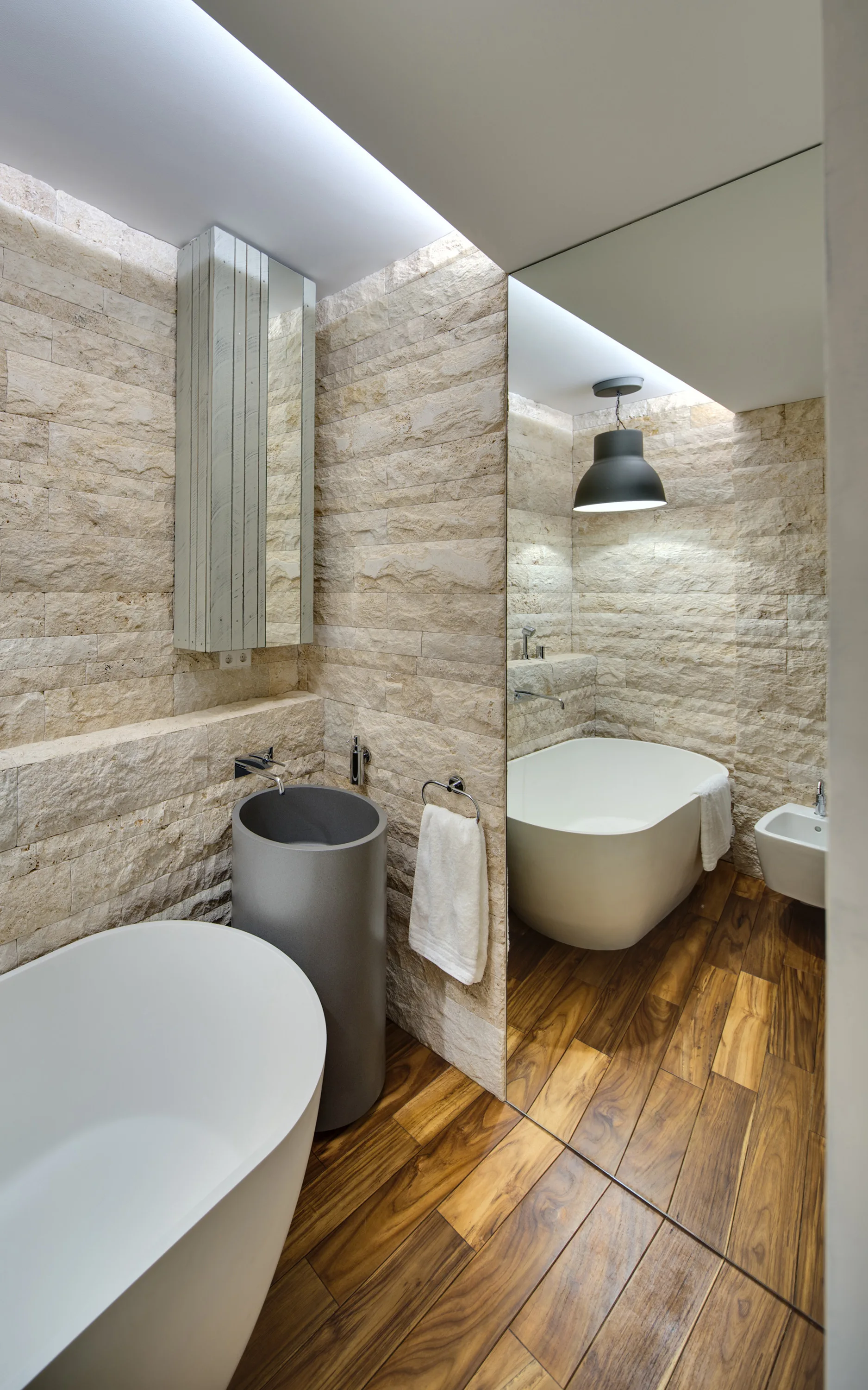 A modern bathroom features textured stacked stone walls, warm wood flooring, a white freestanding bathtub, and a dark gray pedestal sink.