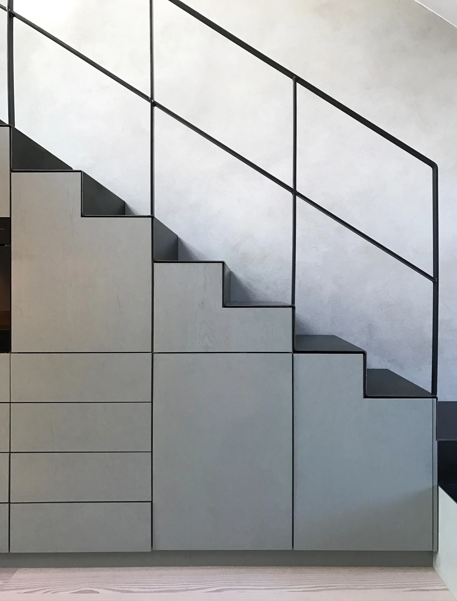A modern staircase features grey built-in cabinets designed to match the step profile and a slim black metal handrail against a light grey wall.
