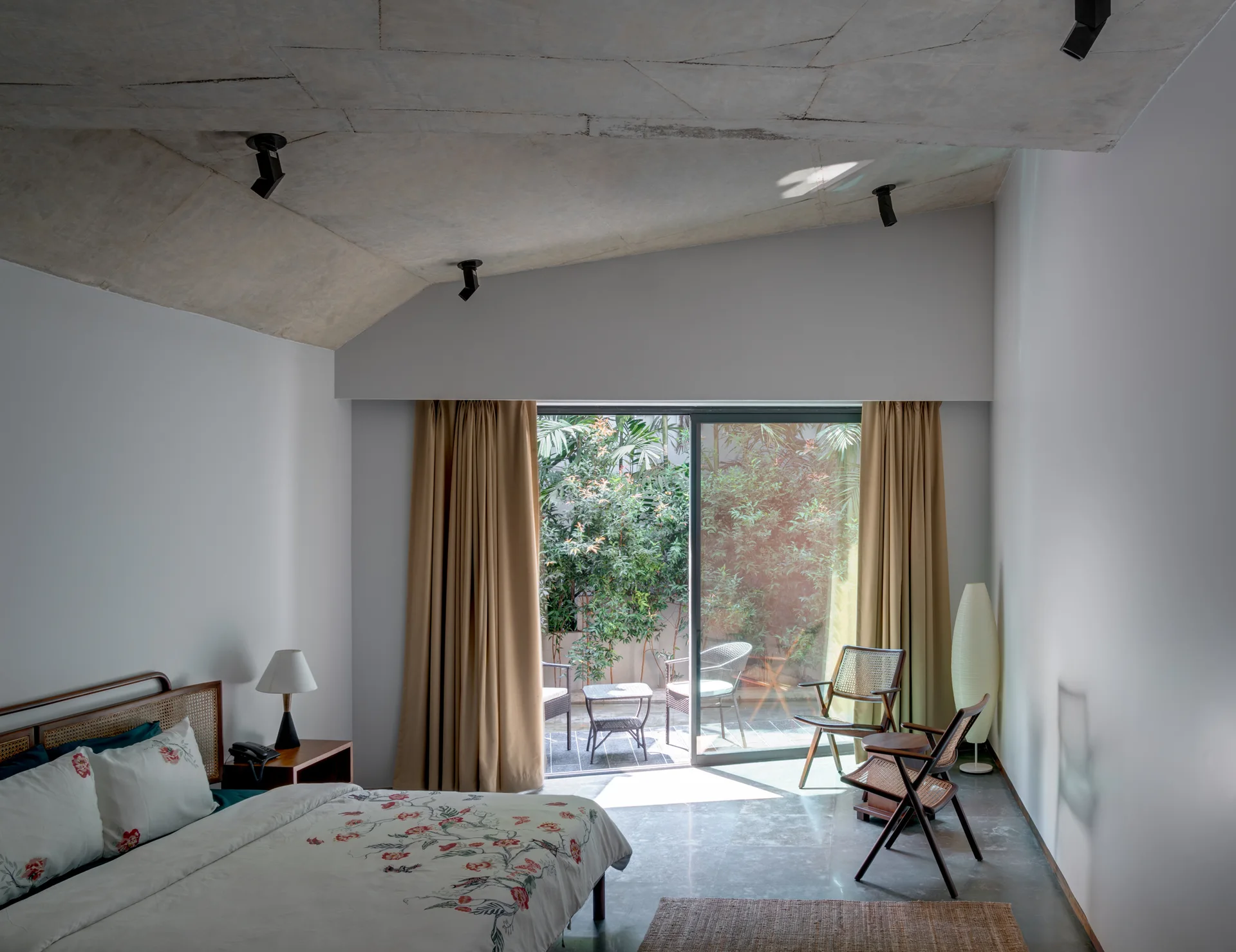A modern bedroom features an angled concrete ceiling, a bed with a floral duvet, and sliding glass doors that open to a lush garden patio.