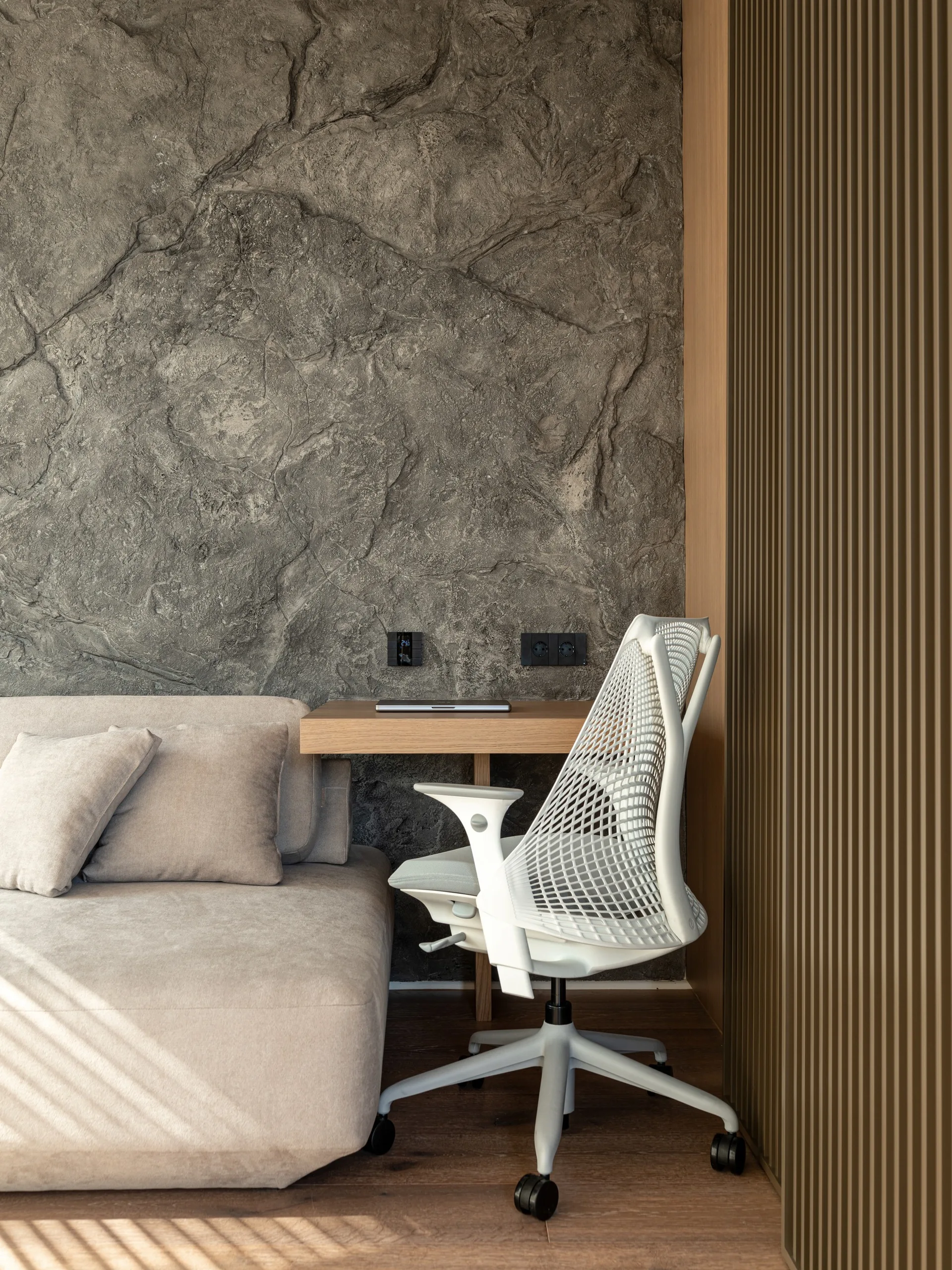 A compact home office setup features a wooden floating desk, a white mesh ergonomic chair, and a rough stone-textured accent wall next to a beige sofa.
