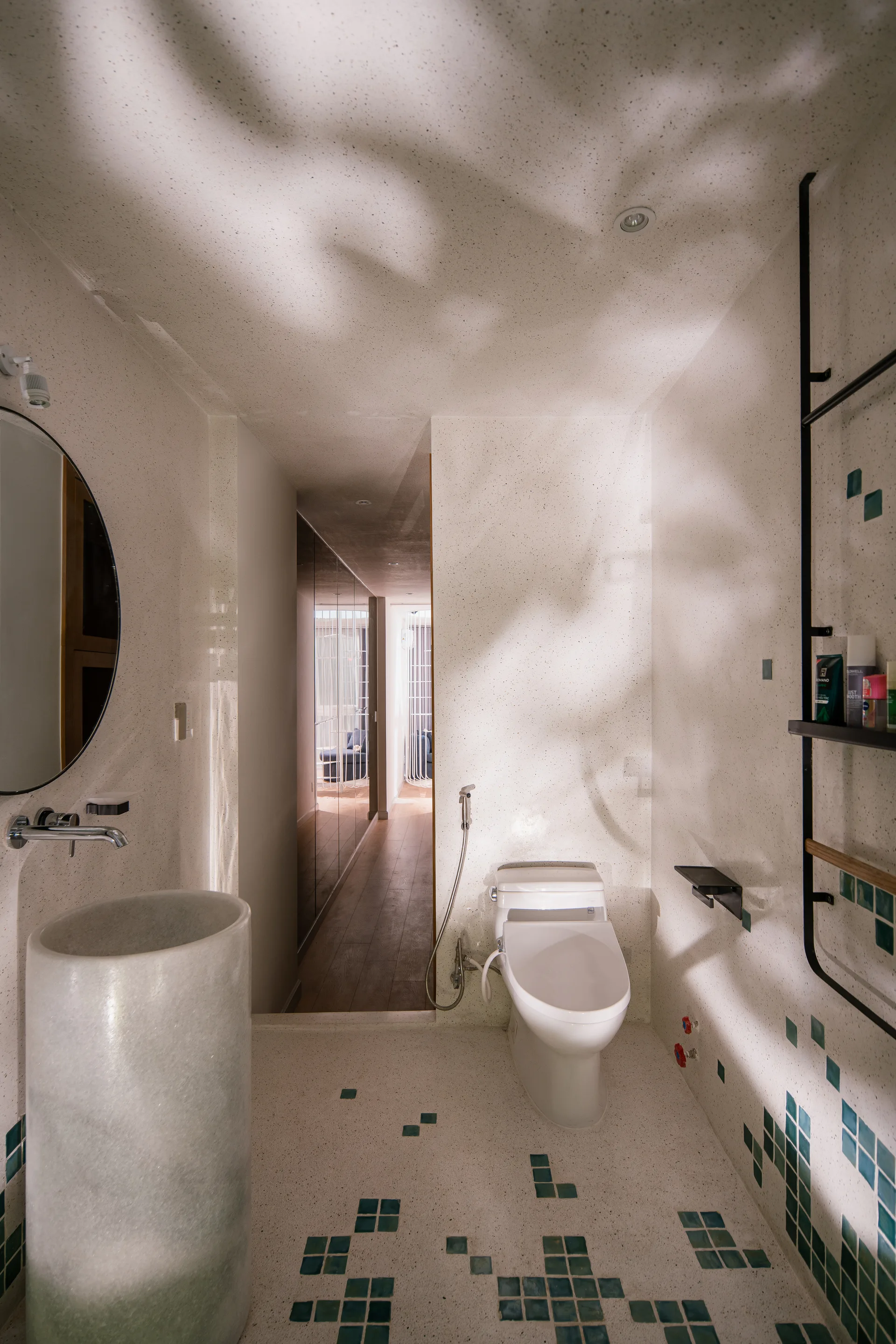 A modern bathroom featuring speckled terrazzo walls and floor, a green-faded cylindrical pedestal sink, a white toilet, and scattered green mosaic tiles.