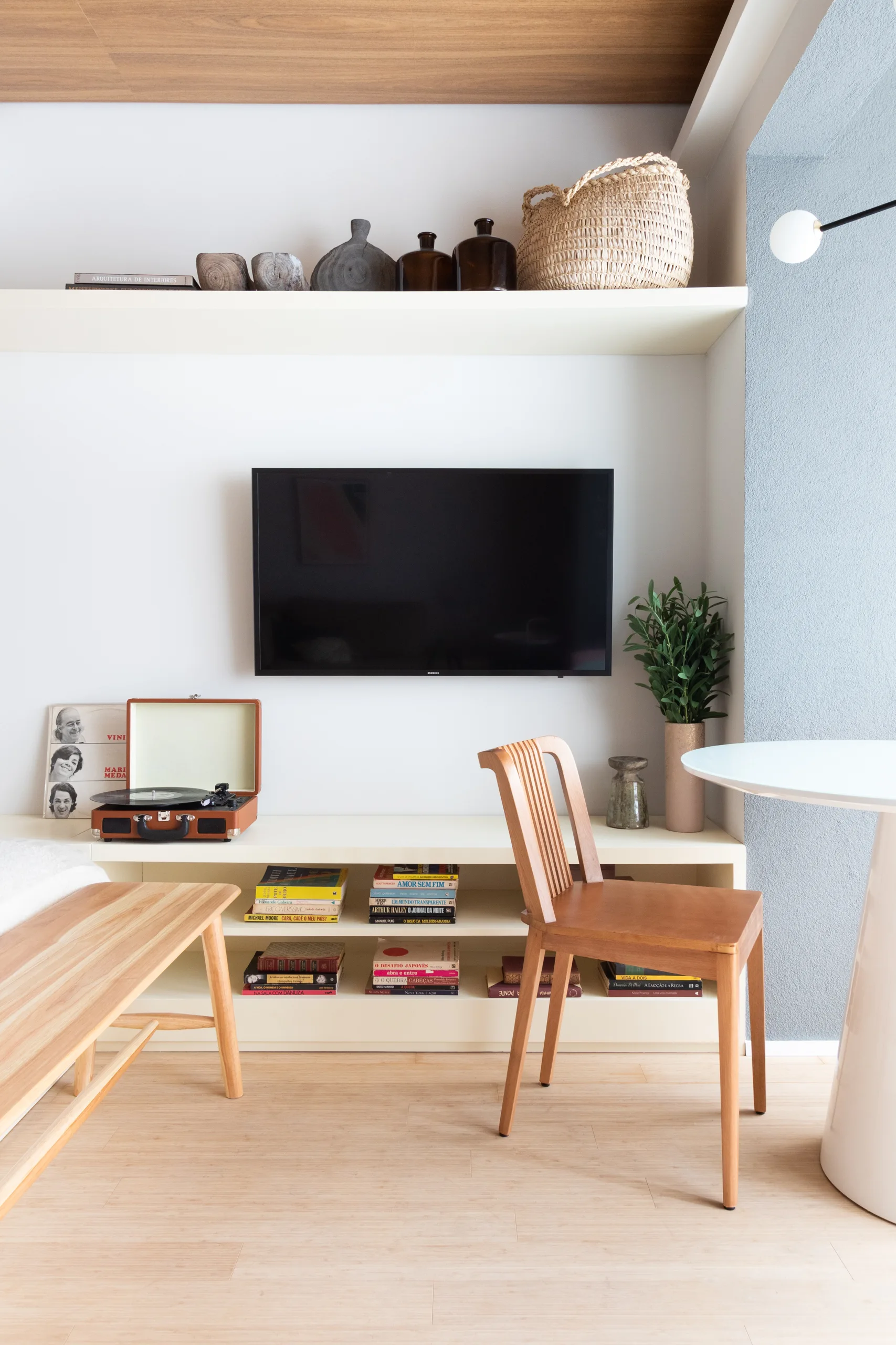 A wall-mounted television is framed by white open shelves displaying a record player, books, a woven basket, and a wooden chair nearby.