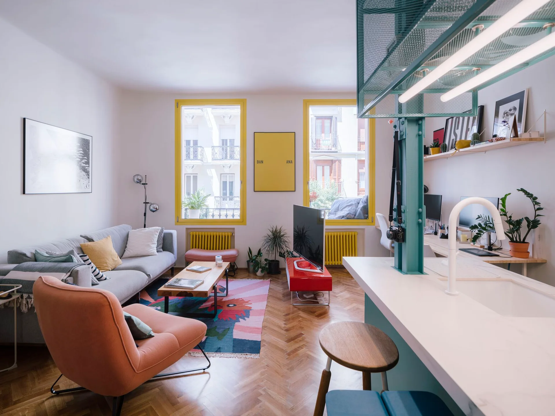 An open living room features a grey sofa, orange armchair, and colorful rug set against herringbone floors and bright yellow-trimmed windows.