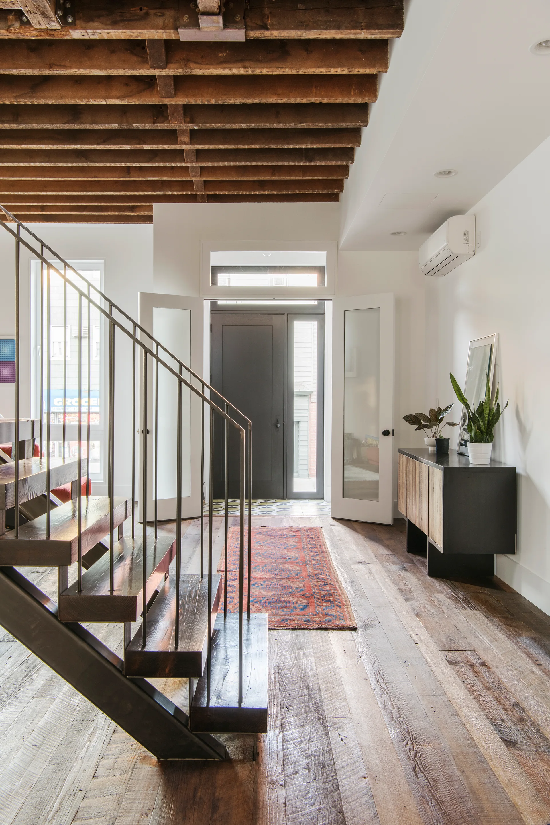 An airy entryway with an open wood and metal staircase, exposed wood ceiling joists, and a runner rug leading to a dark front door.