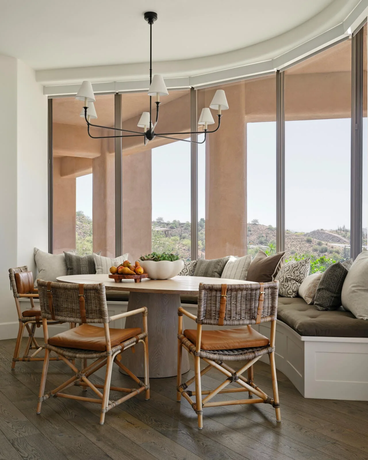 A breakfast nook features a round wooden dining table, woven rattan chairs, and a curved built-in bench set against windows looking out at a desert landscape.