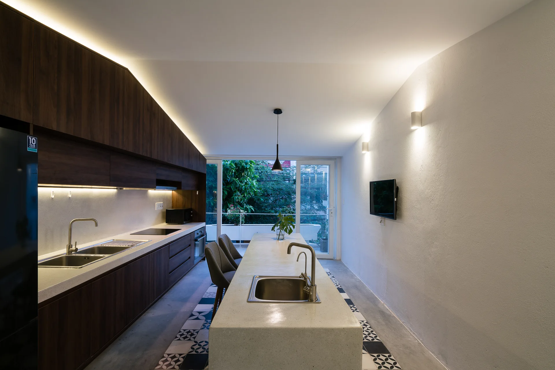 A modern galley kitchen featuring dark wood cabinets, a long concrete island with a sink, and a geometric tiled floor strip.