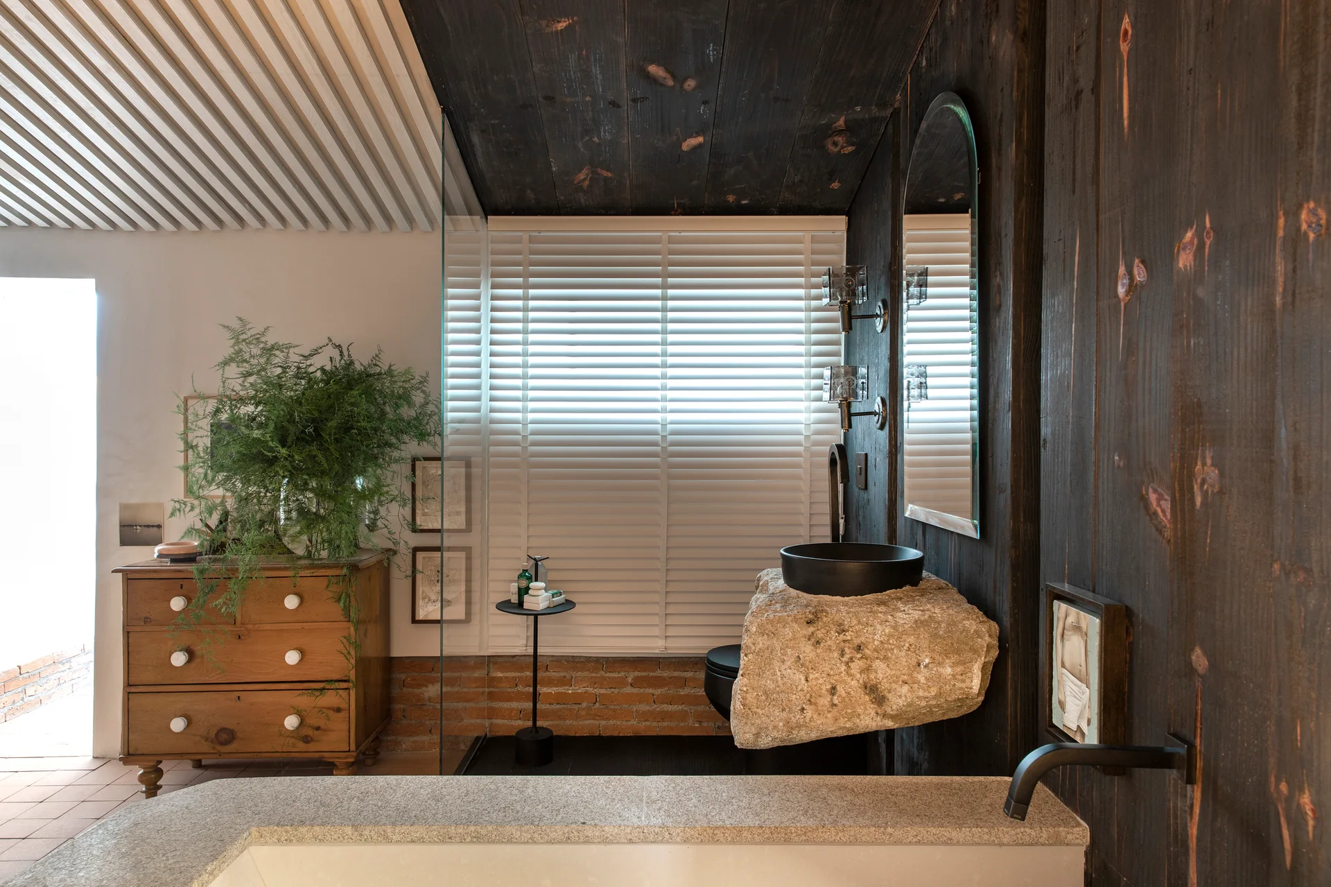 A rustic bathroom features a black vessel sink resting on a rough-hewn floating stone vanity against dark wood-paneled walls.