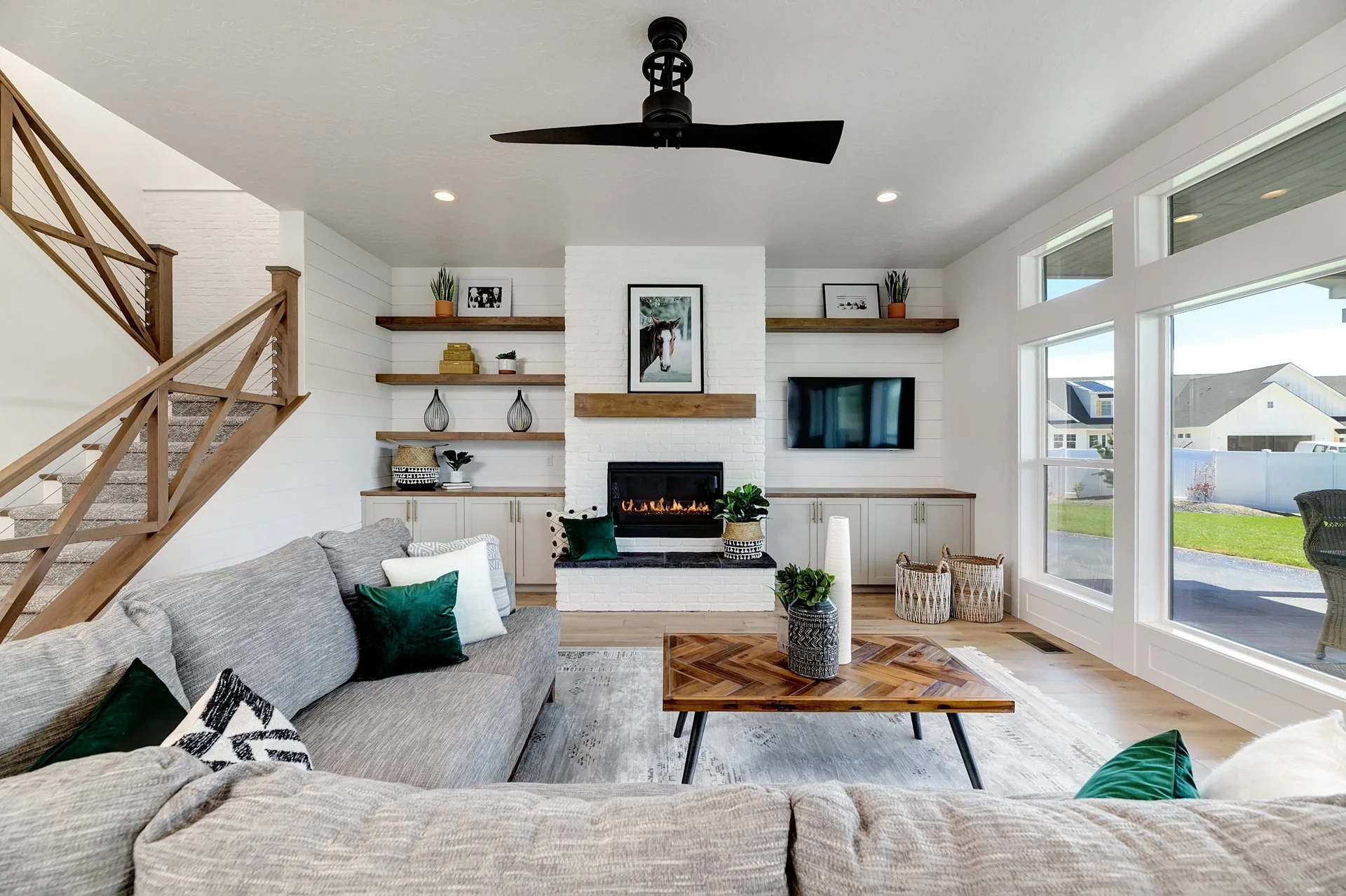 A modern living room features a white brick fireplace with a wood mantel, flanking shelves, a grey sectional sofa, and a chevron coffee table.