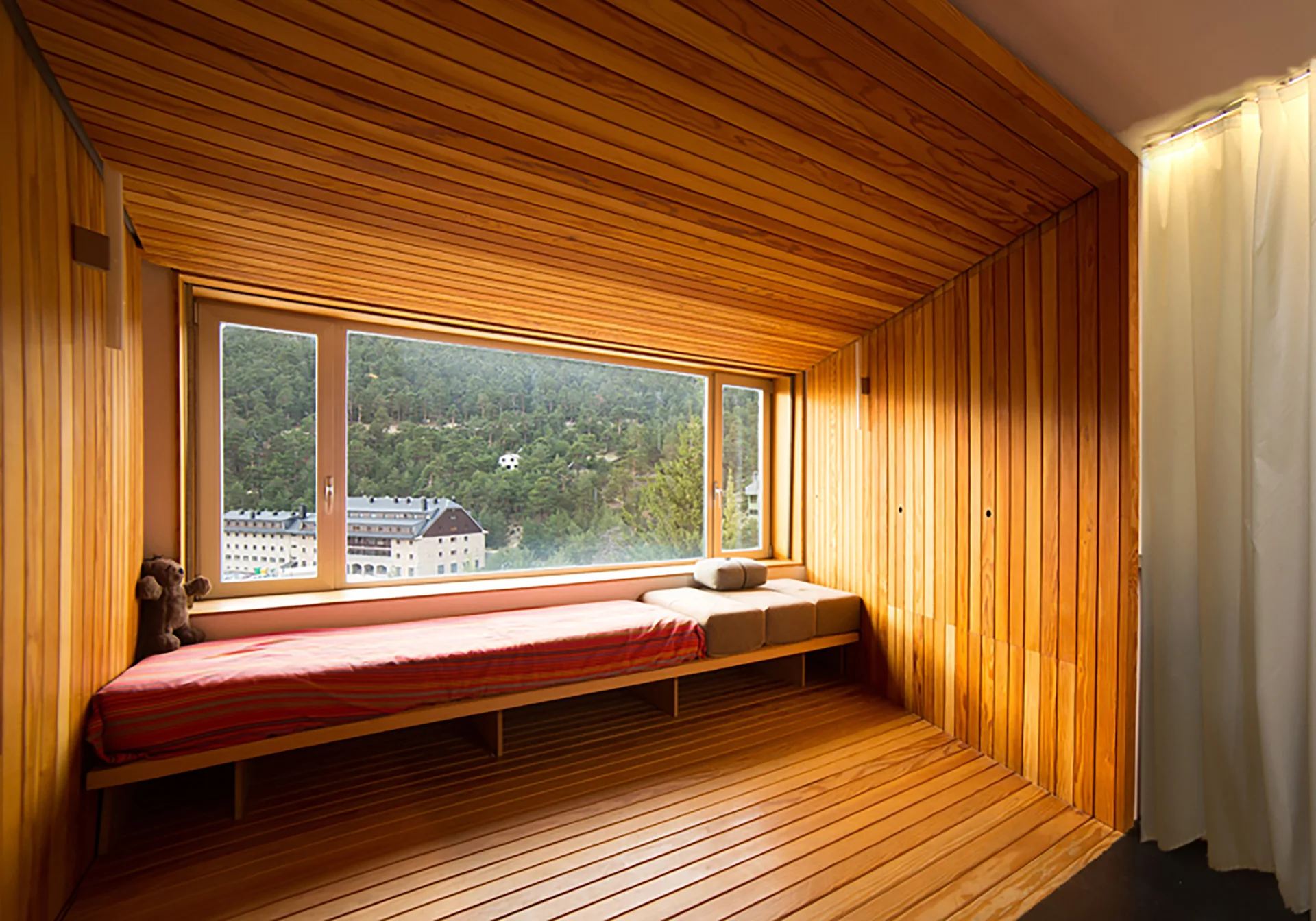 A minimal wood-clad cabin bedroom featuring a built-in daybed beneath a large window showing a forested mountain landscape.