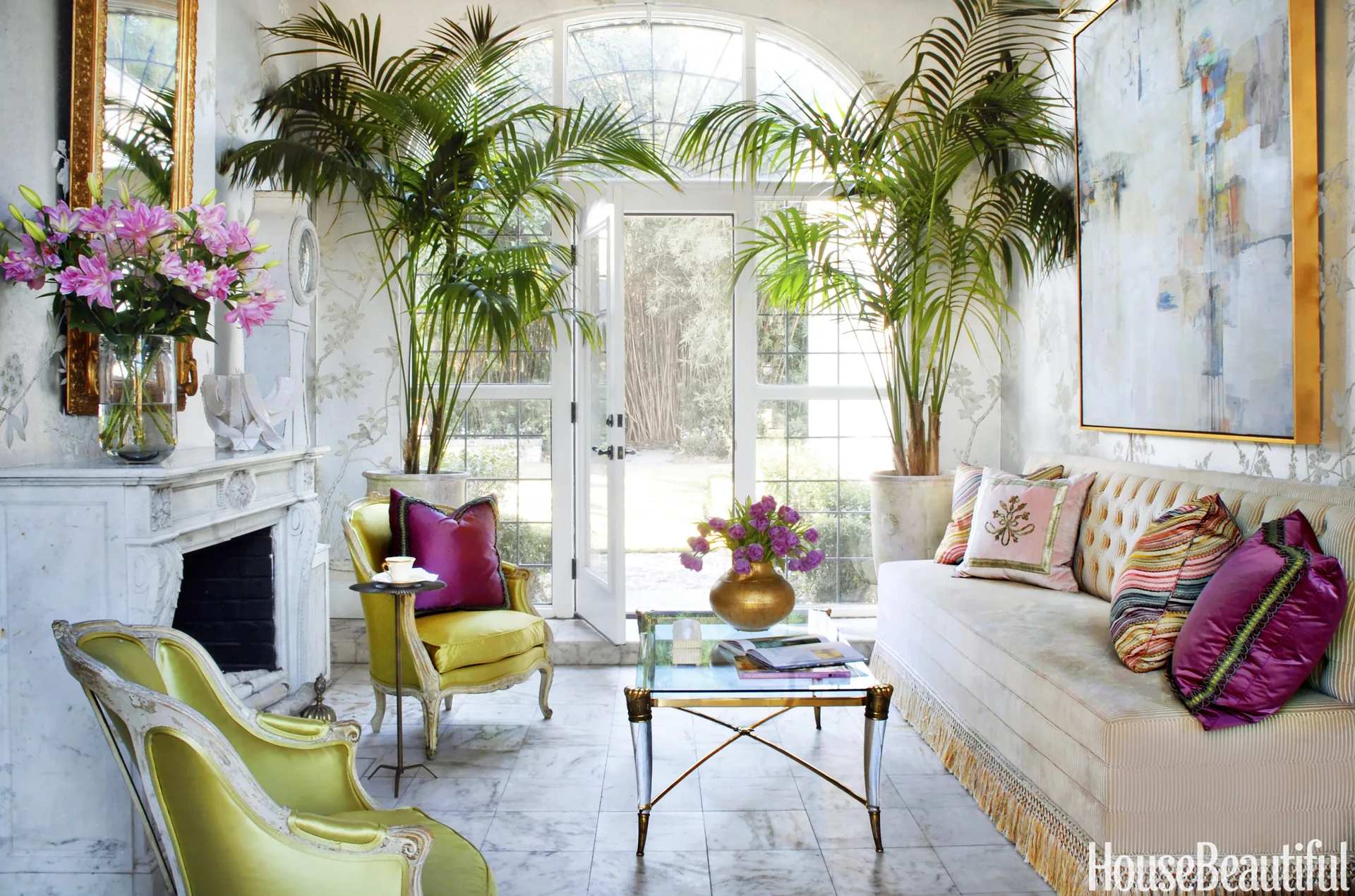 A bright sunroom features a tufted cream sofa, two light green armchairs, a marble fireplace, and tall potted palms beside an arched glass door.