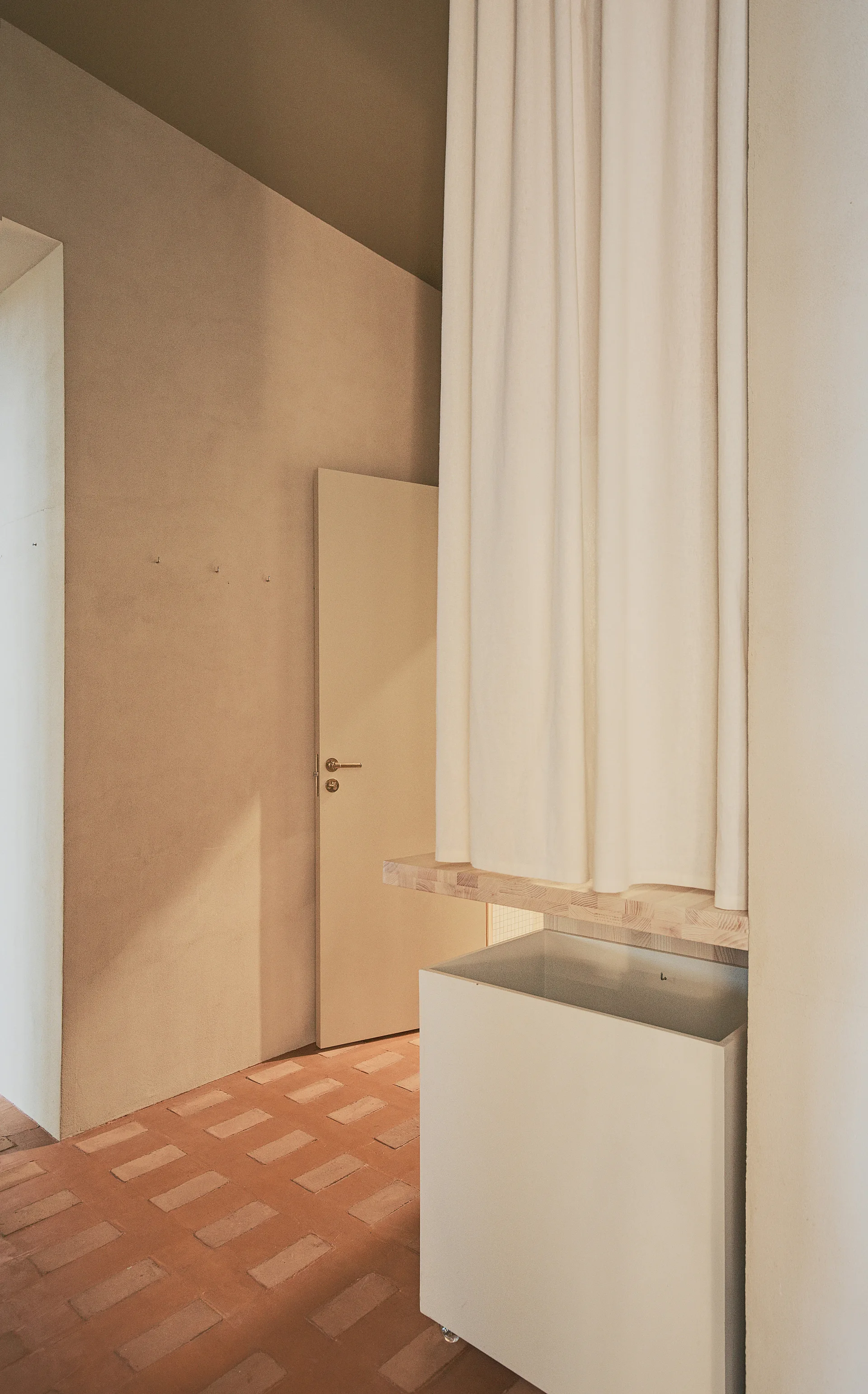 A minimalist hallway with a smooth beige wall, a white curtain partition over a wood shelf, and terracotta brick flooring.