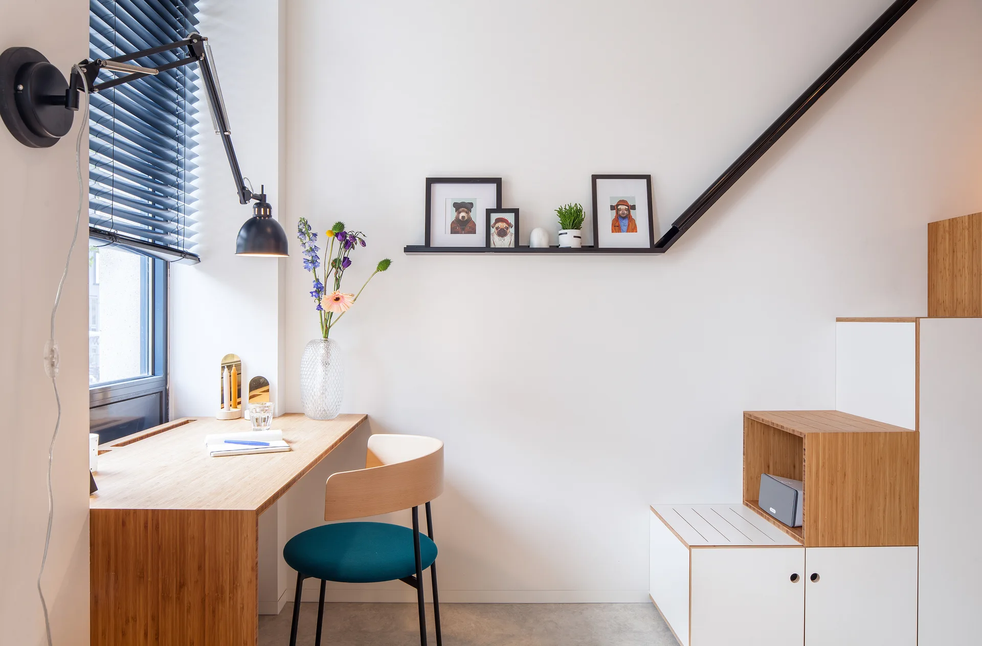 A light wood desk with a teal cushioned chair sits next to a window with dark blinds, beneath a black wall-mounted lamp and floating shelf.