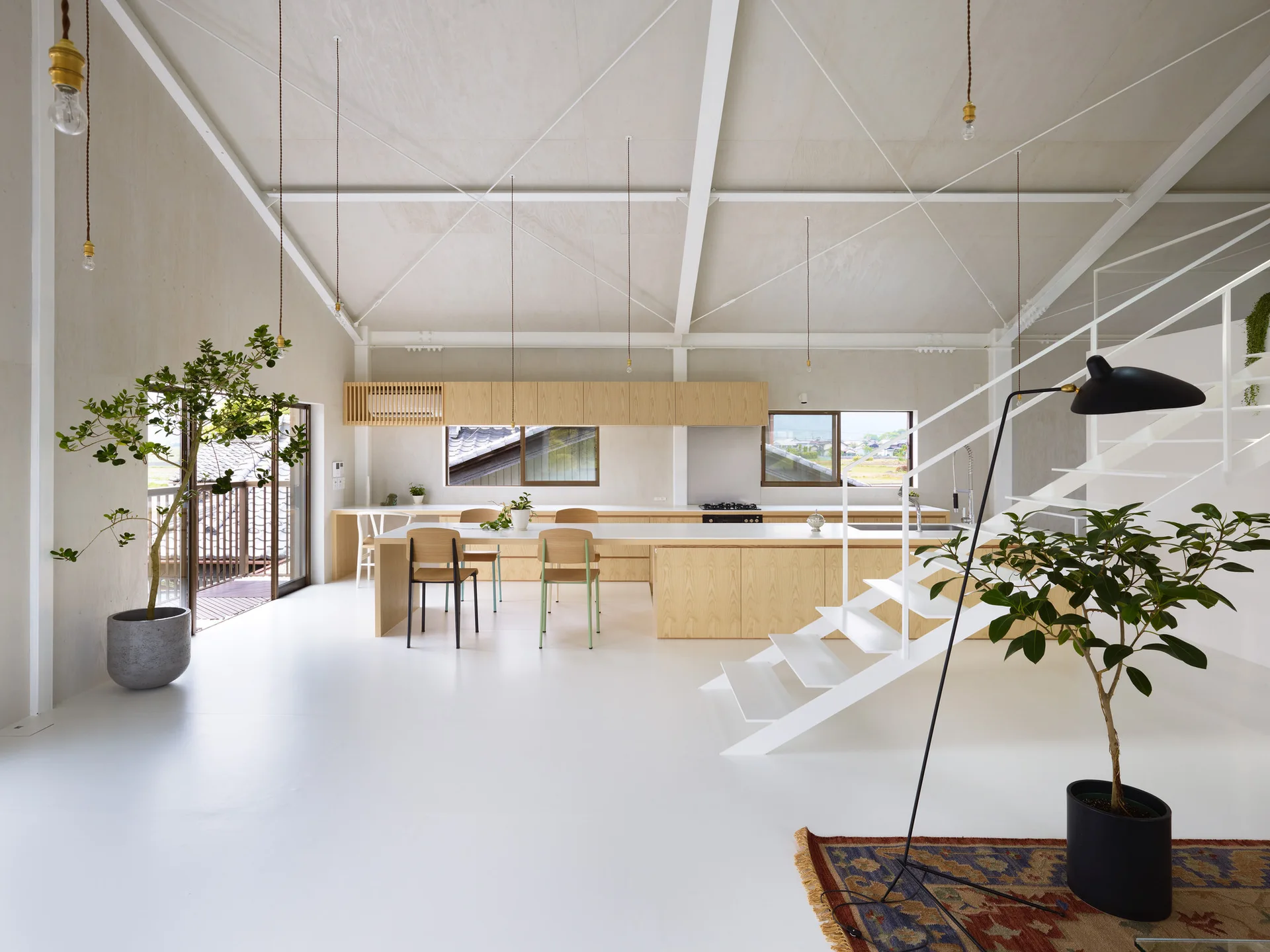 A minimalist open-plan kitchen features a long light wood island, a white steel staircase, and exposed structural ceiling trusses.