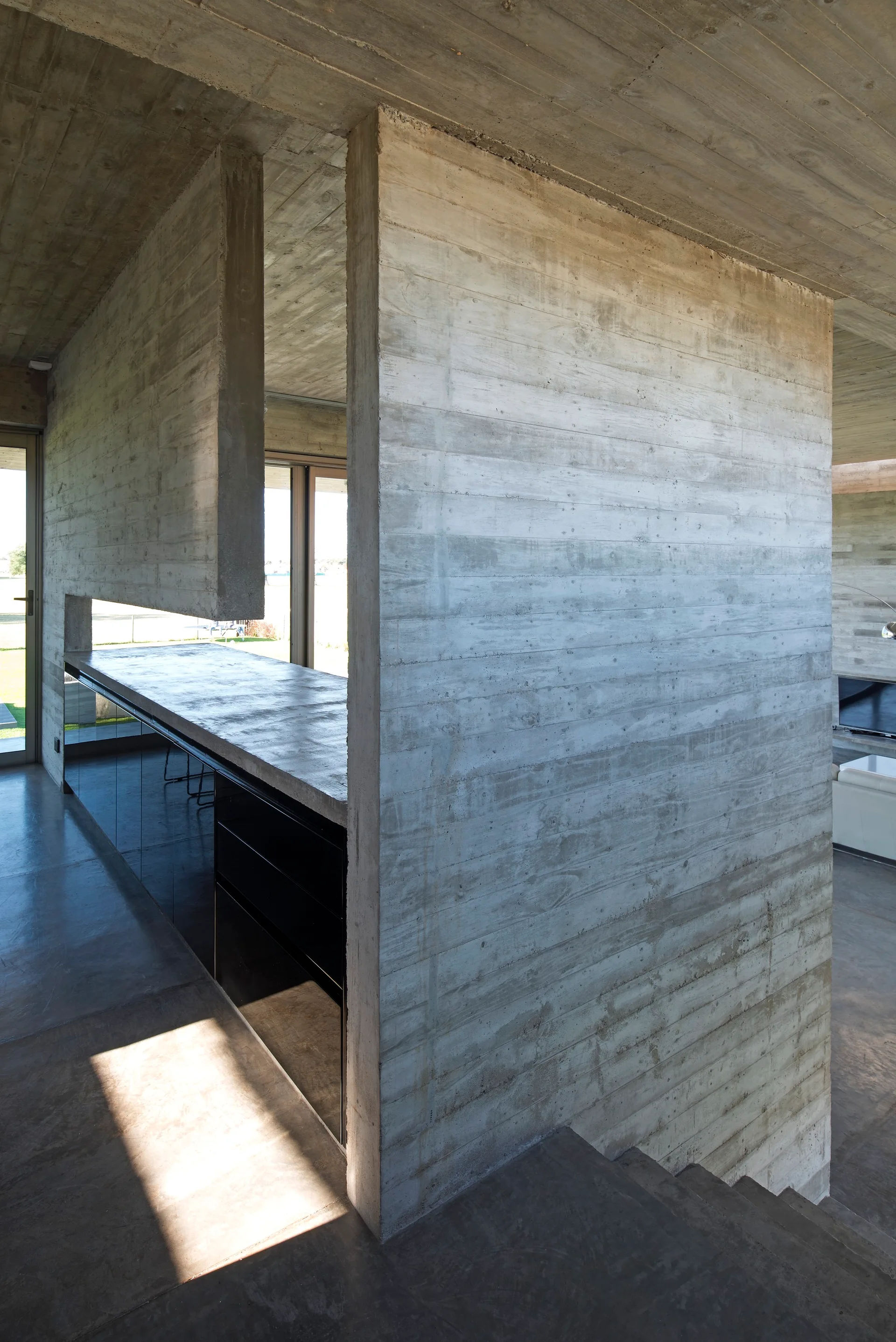 A minimalist kitchen features raw board-formed concrete walls, concrete countertops, and reflective black cabinetry next to a structural partition.