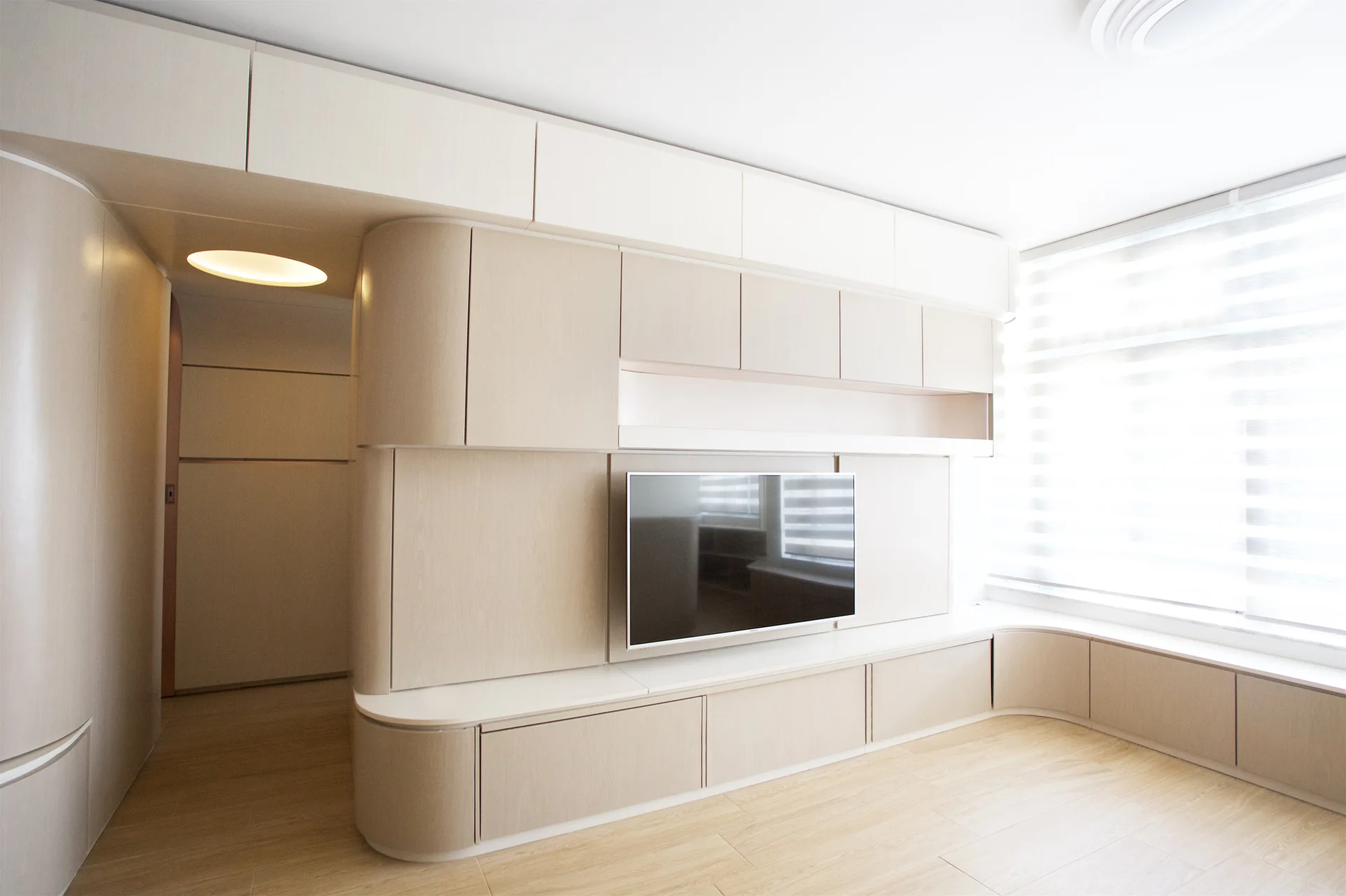 A modern living room features light wood custom cabinetry with rounded edges, a wall-mounted TV, and an integrated window bench.