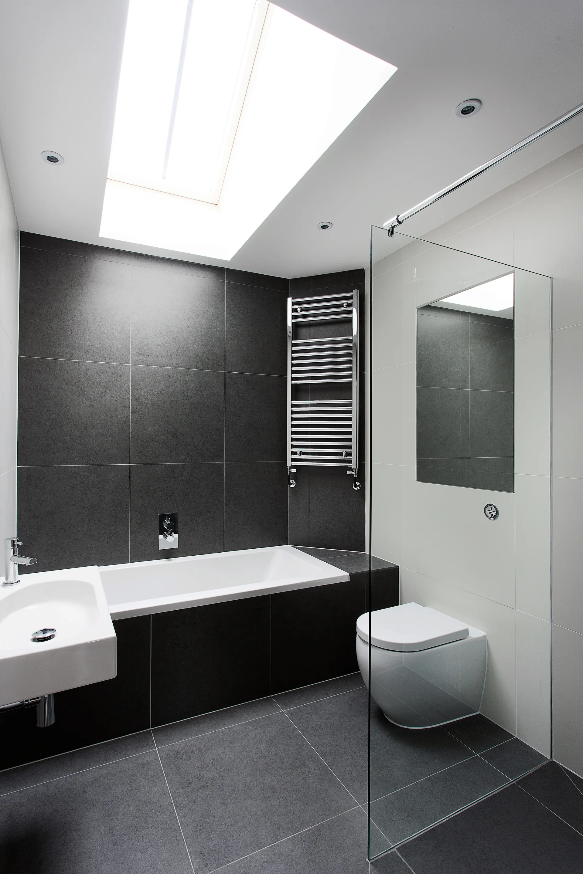 A modern bathroom with black tiled walls and floor features a white bathtub under a skylight, a floating toilet, and a glass partition.
