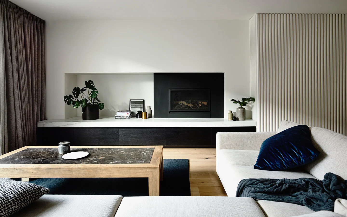 A modern living room features a cream sofa, a marble-topped coffee table, and a black fireplace surrounded by a white wall and vertical wood slats.