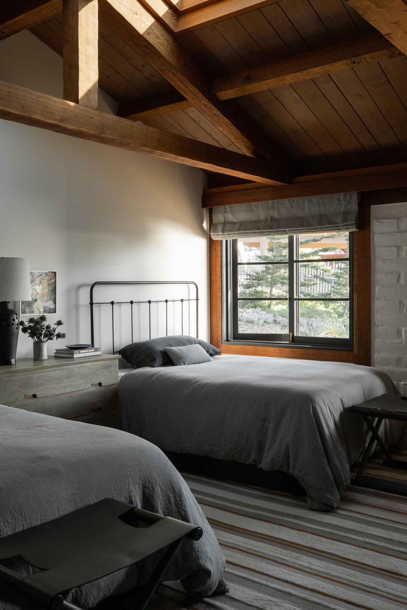 A rustic bedroom features two beds with charcoal gray linen bedding, black iron headboards, and a high ceiling with exposed wooden beams.