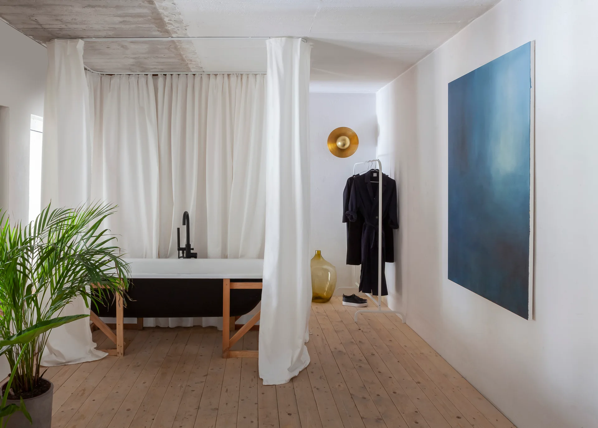A black freestanding bathtub enclosed by white fabric curtains sits on light wood flooring next to a large blue canvas painting and a green plant.