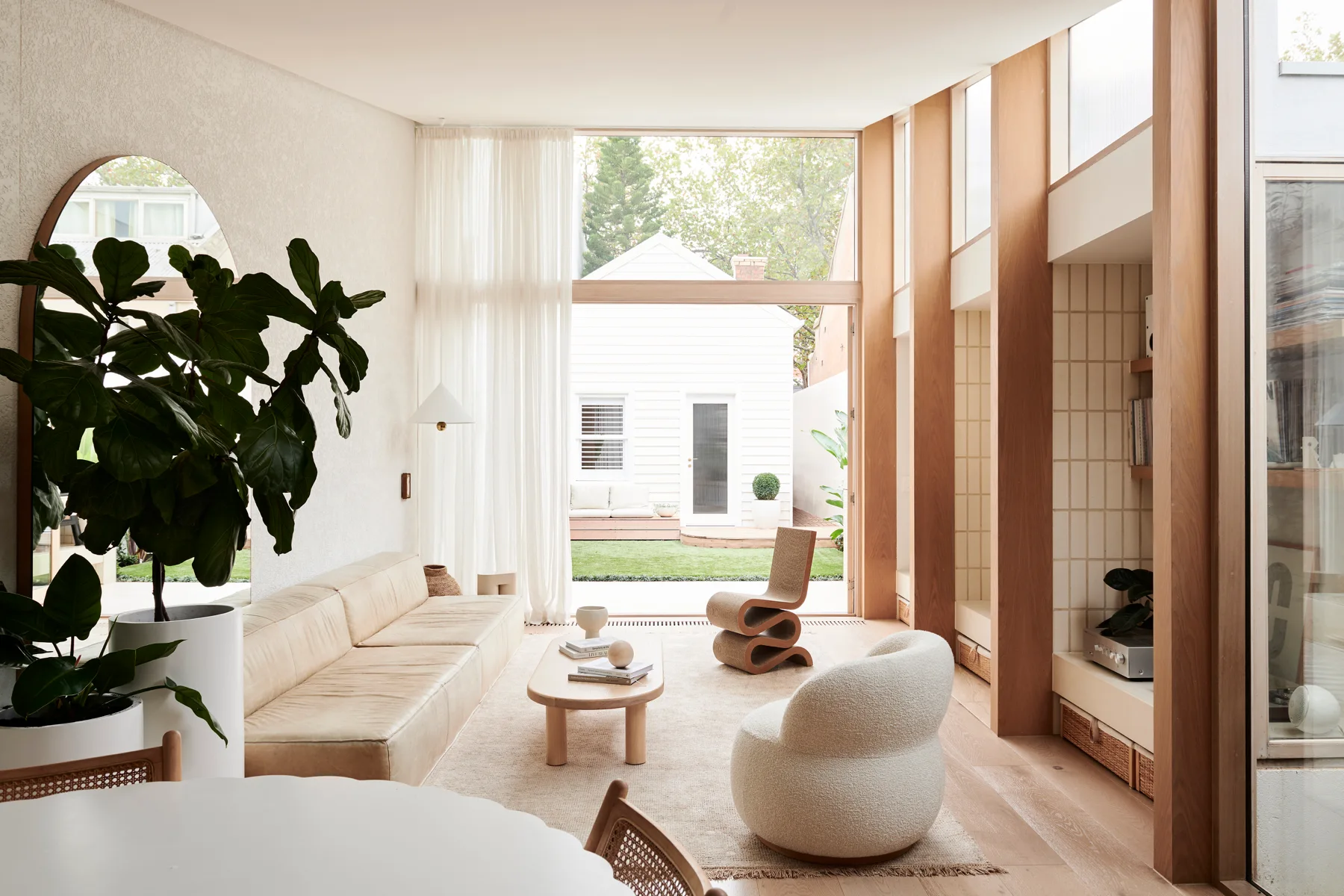 A modern living room features a beige leather sofa, a textured white boucle armchair, a large arched mirror, and floor-to-ceiling windows.