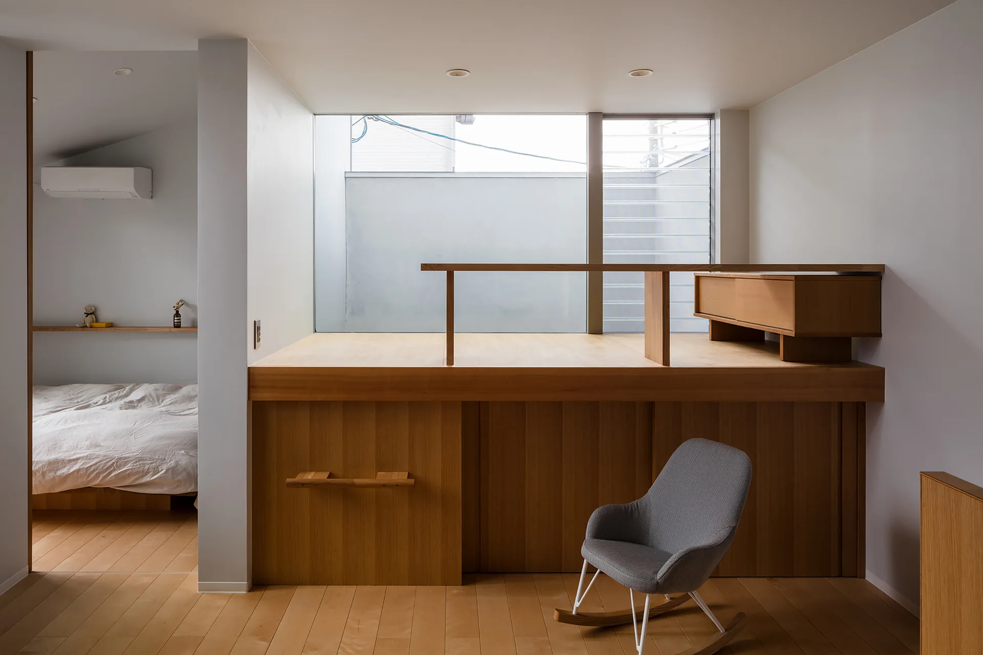A minimalist bedroom featuring a raised wooden platform under a large window, a grey rocking chair, and a separate bed area in the background.