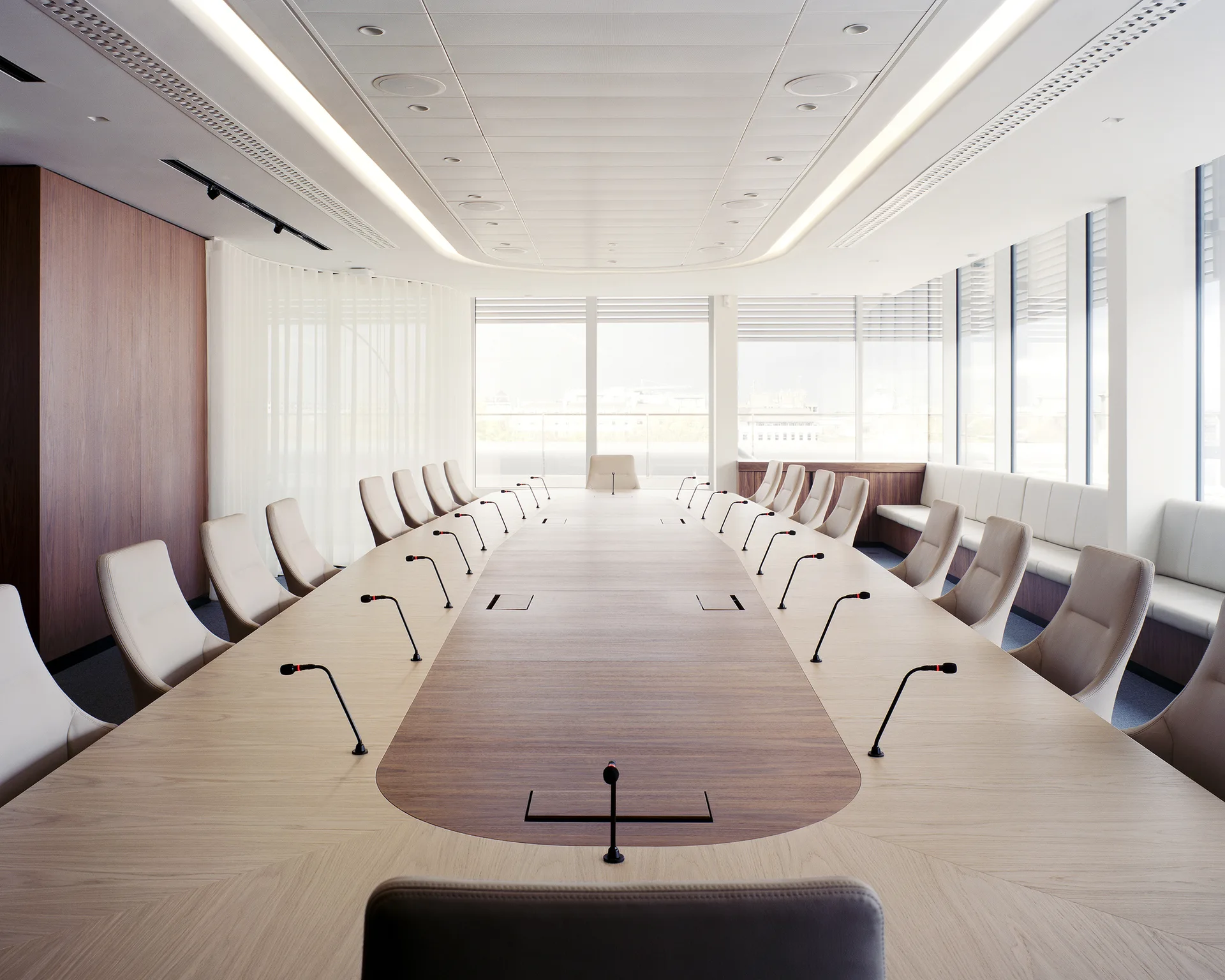 A spacious boardroom features a long oval wooden table with gooseneck microphones, surrounded by cream high-back chairs under recessed ceiling lights.