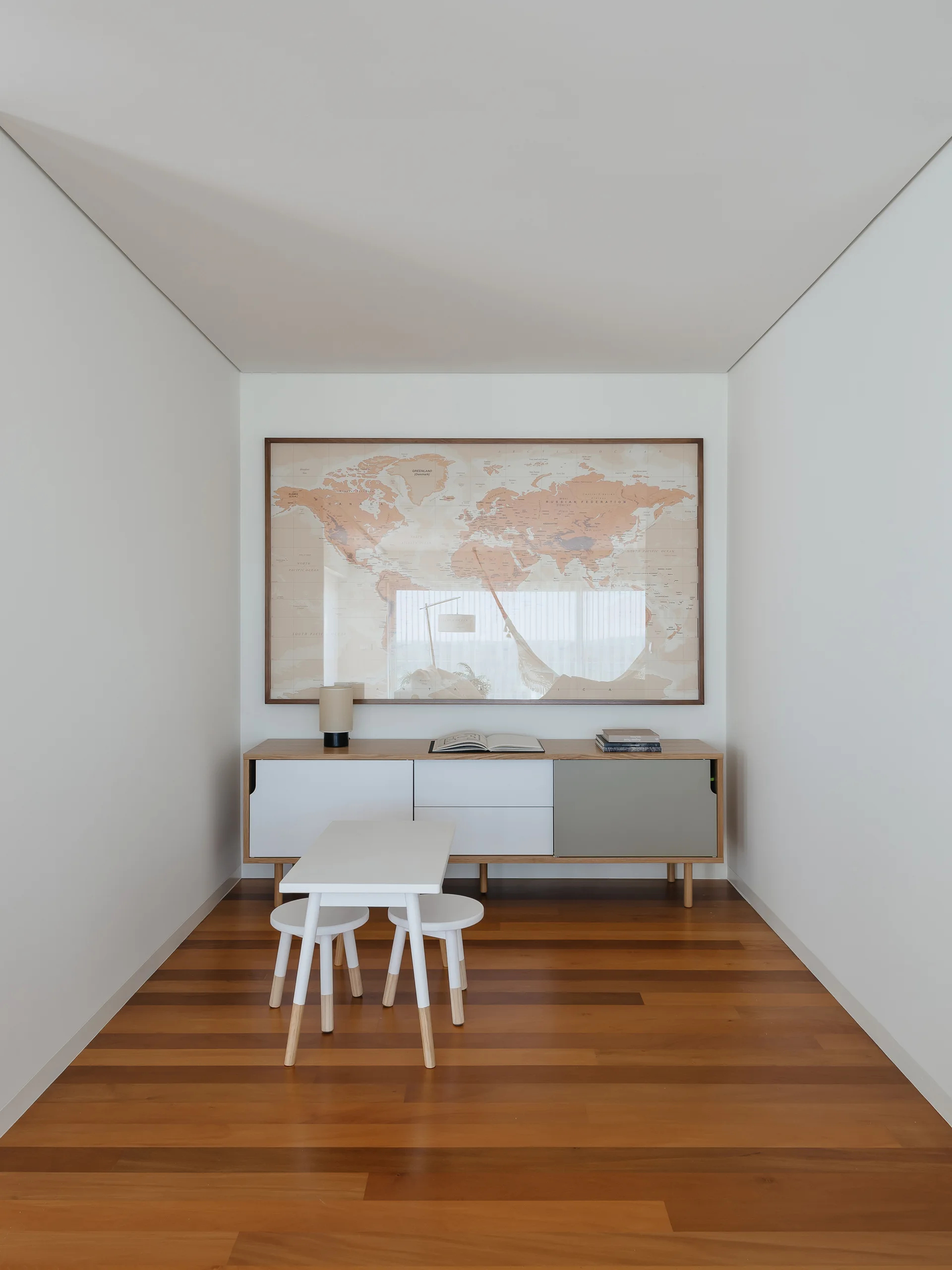 A minimalist white alcove features a low wood sideboard under a framed world map, with a small white children's table and stools in front.