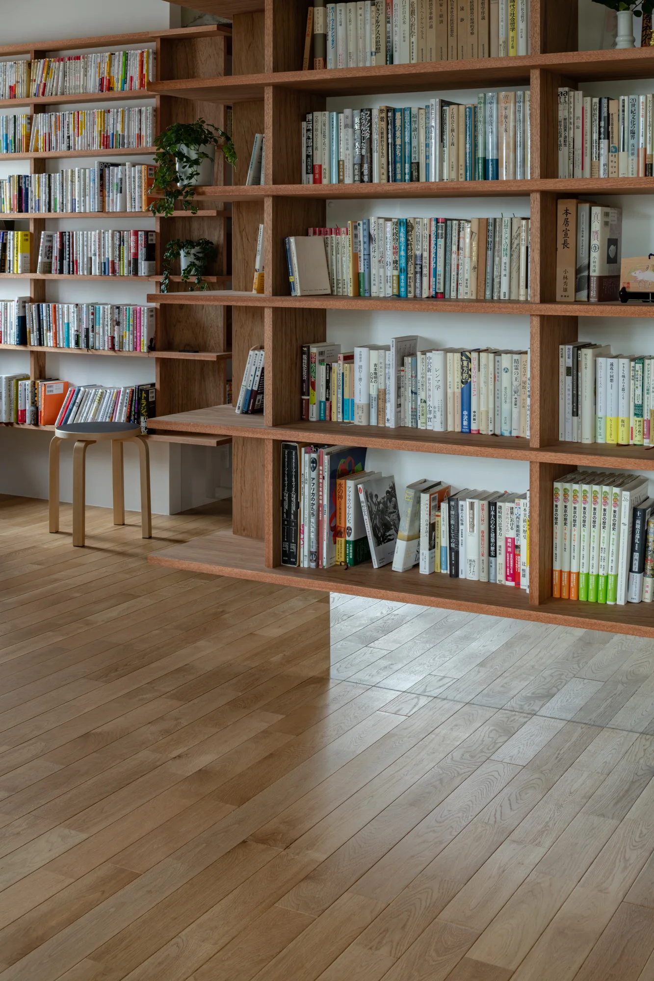 A large wall-mounted wooden bookshelf filled with books is set against a white wall above light-toned hardwood floors, with a small stool on the left.