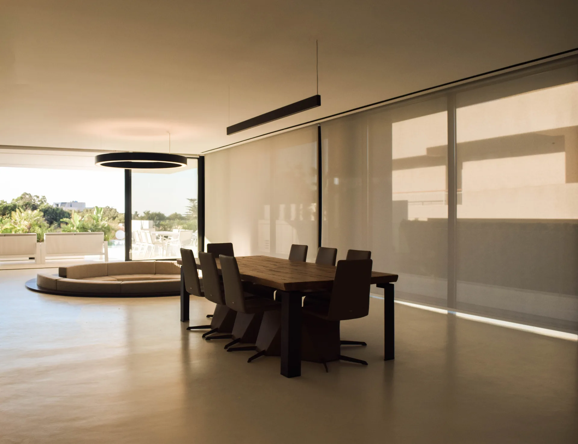 A long wood dining table and dark chairs sit in a minimalist room with large roller shades and a circular sunken lounge in the background.