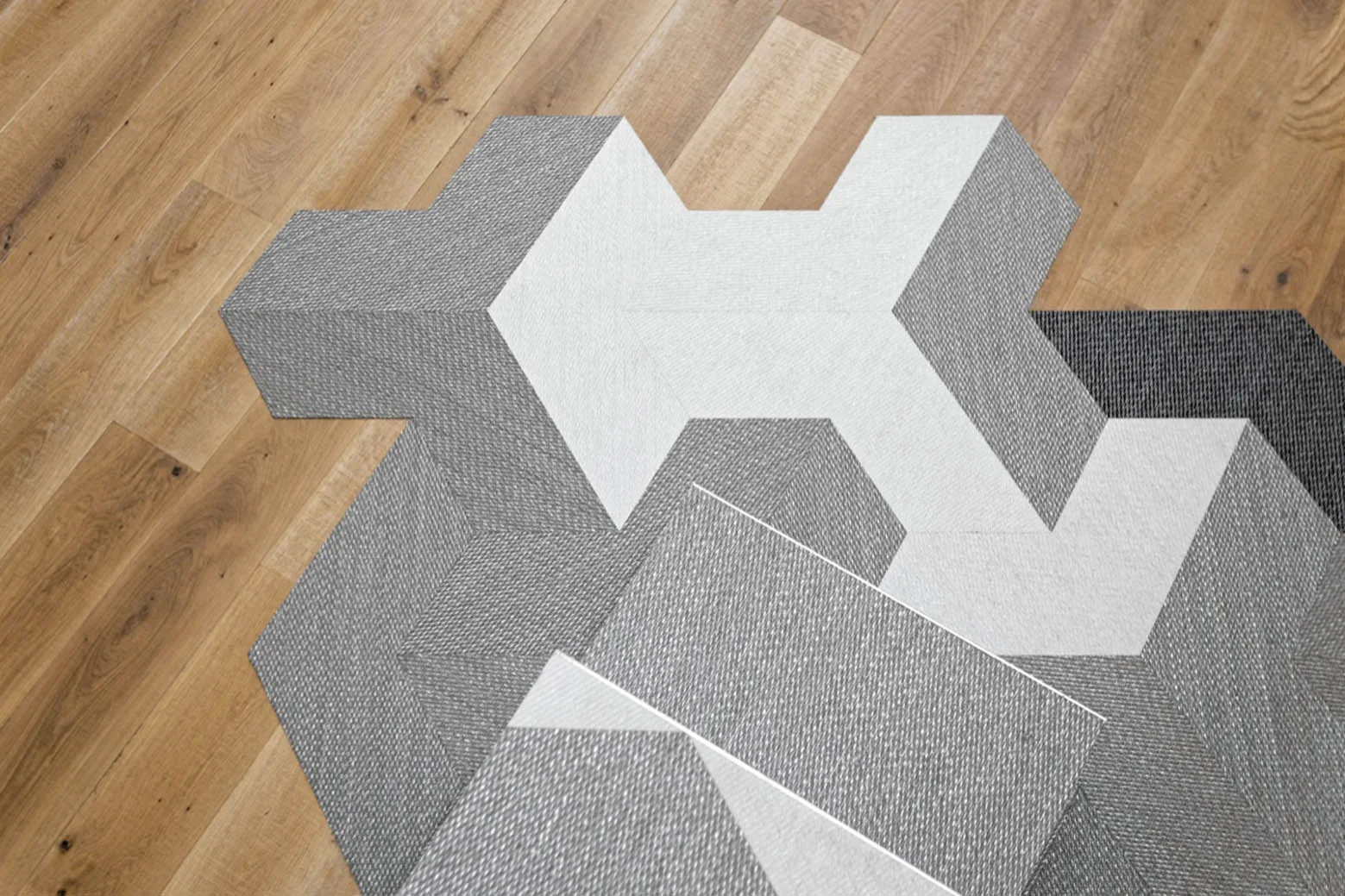 Geometric grey and white modular carpet tiles with a woven texture are arranged on a light oak wood plank floor.