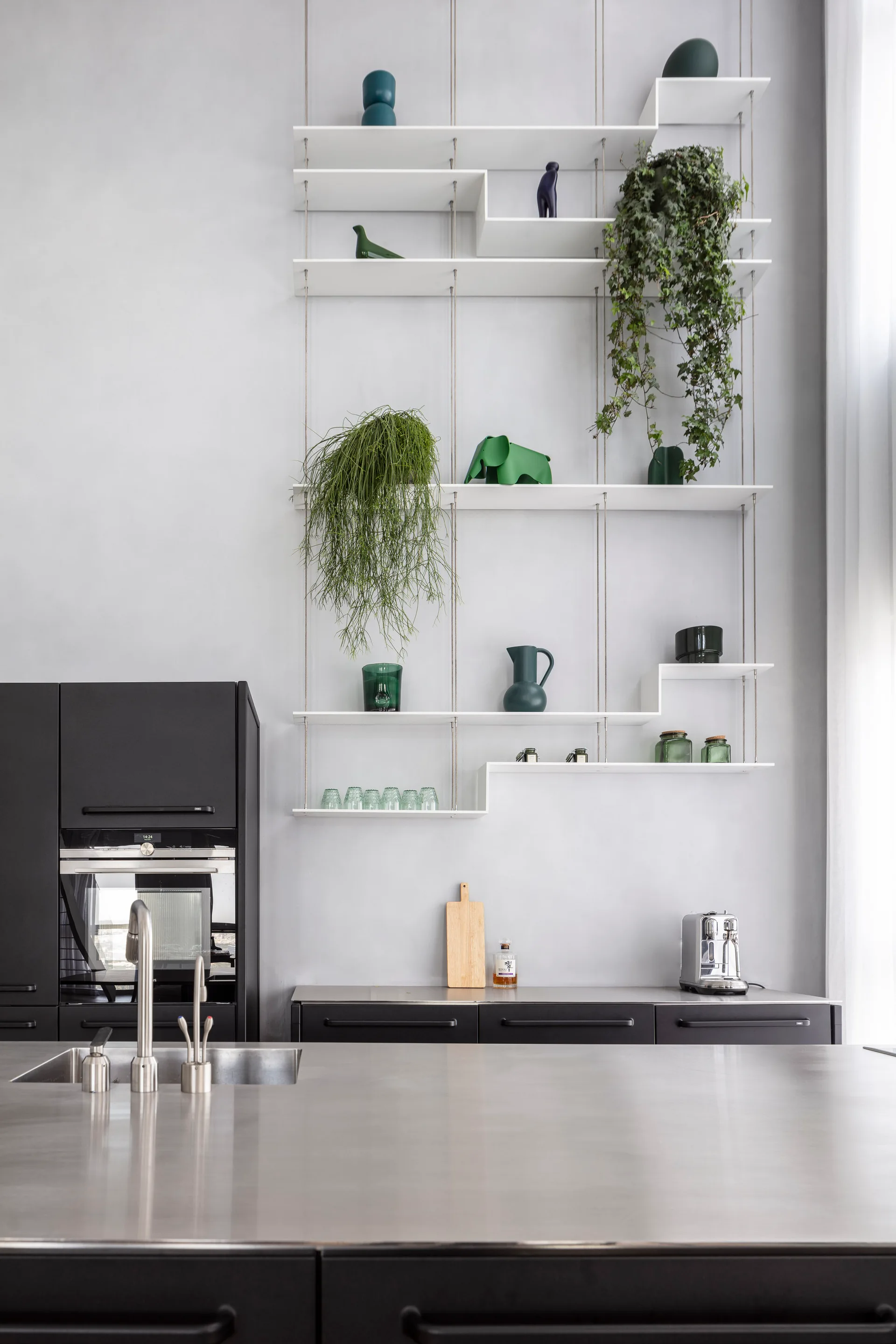 A modern kitchen with black cabinets, a stainless steel island, and white suspended open shelves displaying green plants and decor.