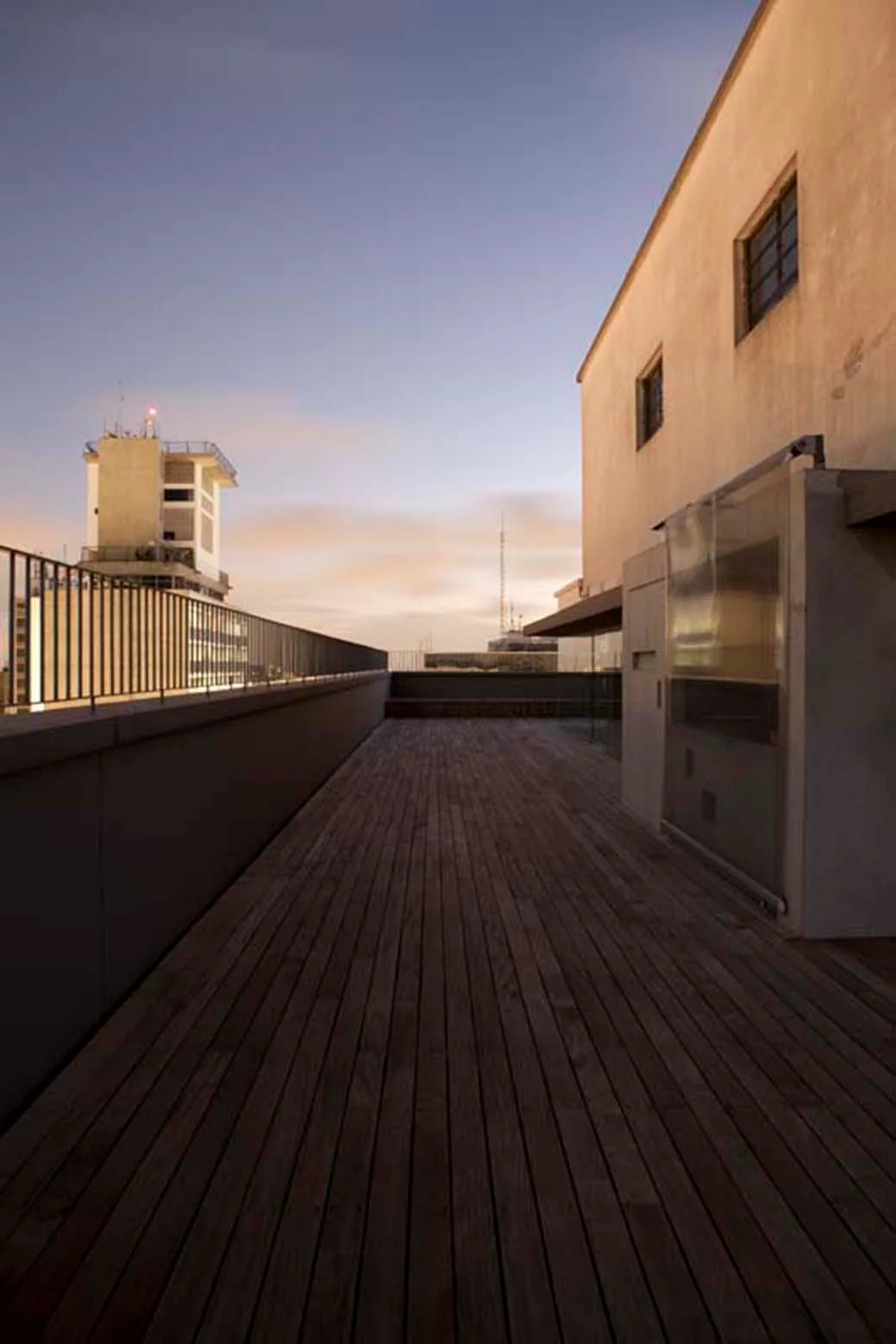 A long wooden plank deck on a rooftop terrace features a metal railing and an adjacent beige building facade under a twilight sky.